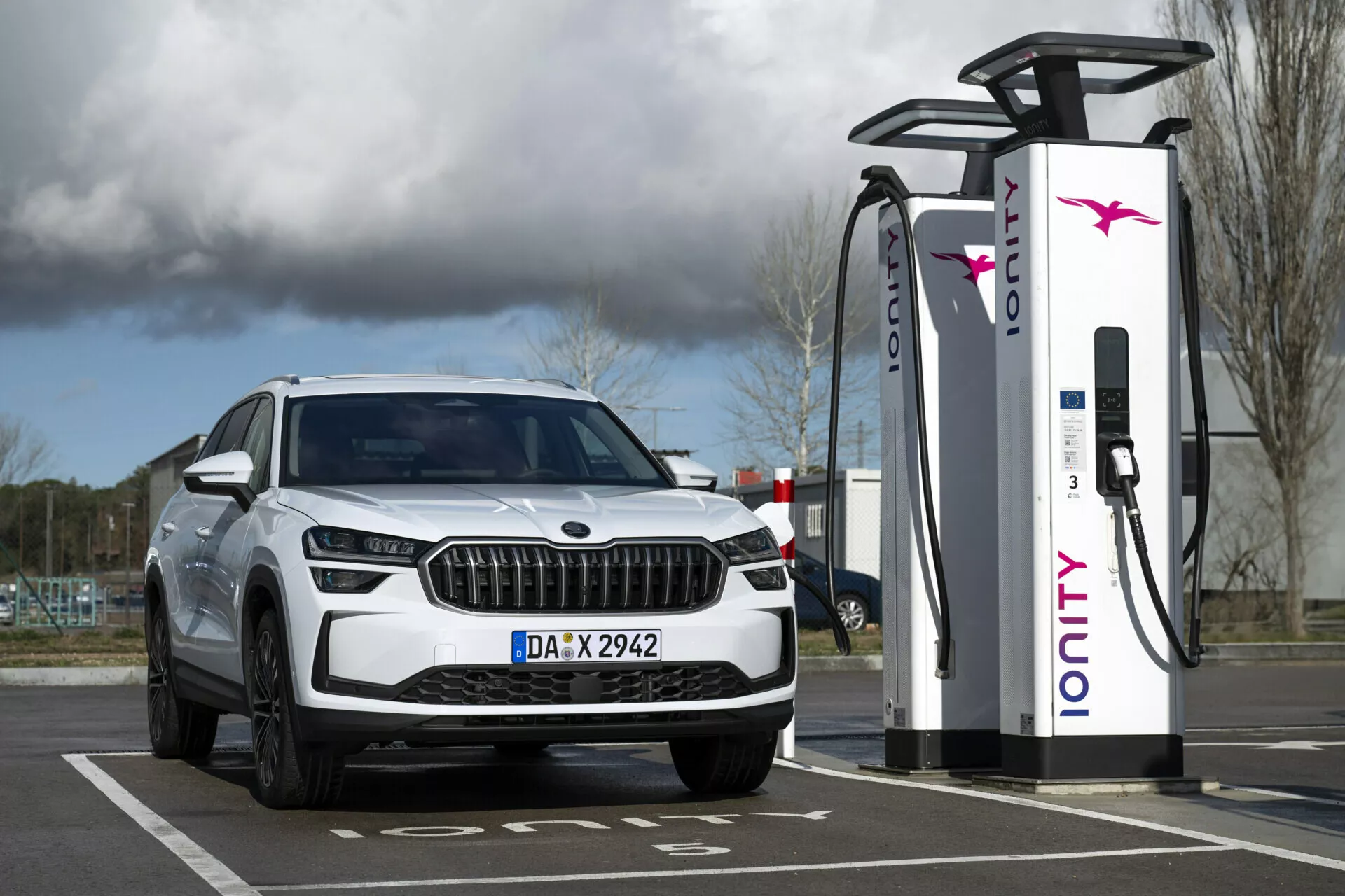 2024 Skoda Kodiaq vit SUV parkerad vid en elektrisk laddningsstation, visar främre kvartalet med LED-strålkastare och dagtidljus, vilket betonar dess elektriska eller hybrida funktioner och lyxig grilldesign.
