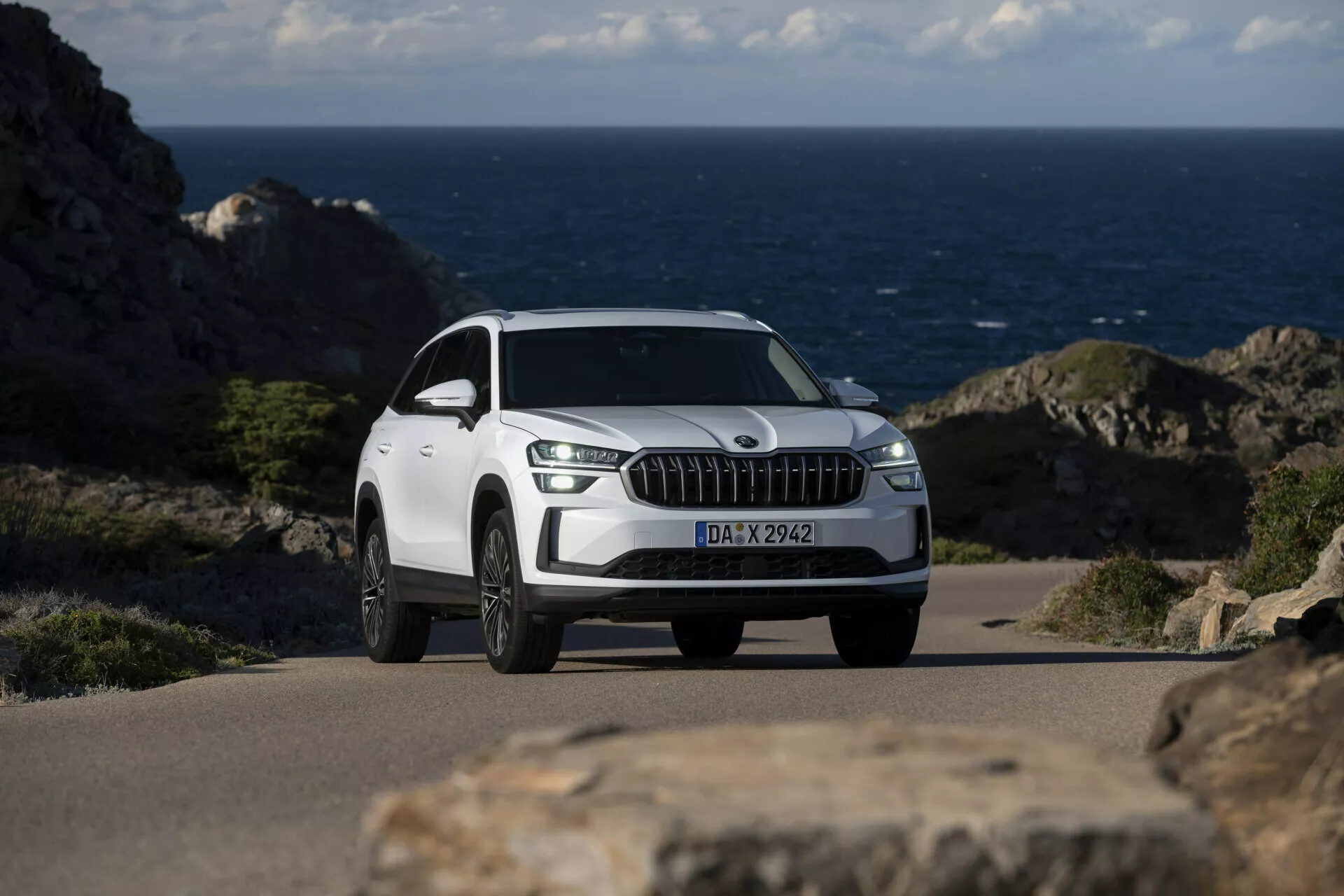 Vit SUV Skoda Kodiaq 2024 parkerad på en kustväg med oceanen i bakgrunden, med en framträdande frontgrill med vertikala kromliknande ribbor, LED-dagsljus, legerade flerspakiga hjul och en detaljerad vy från framsidan i tre fjärdedelar
