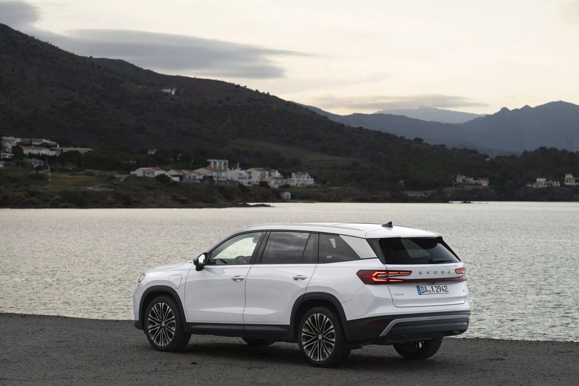 Modern SUV Skoda Kodiaq 2024 parkerad sidledes på en naturskön kustväg, med aerodynamisk design, bakljusremsa och flerspakiga legeringshjul, på en mulen dag med diffust naturligt ljus, med bakgrund av vatten, kullar och byggnader
