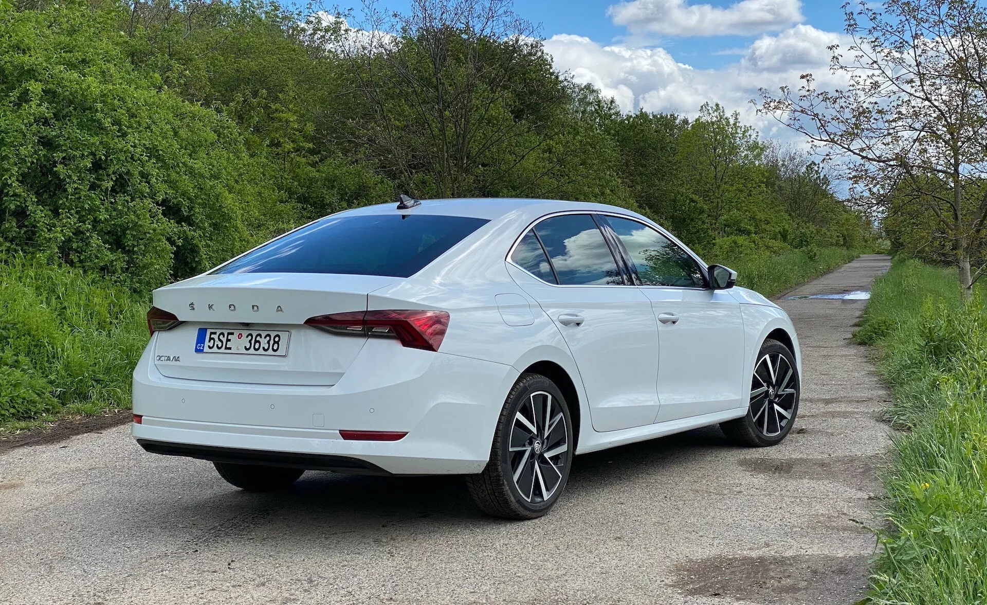 Bakifrån trekvartsvy av en vit Skoda Octavia 2020 sedan parkerad på höger sida av en smal väg, som visar passagerarsidan från marknivå. Bilen fyller det mesta av ramen, och framhäver desiglinjer, bakljus som sträcker sig runt bakluckan, subtil integrerad spoiler, bakre badgering, mångekrade lättmetallfälgar, svagt tonat bakfönster och sidospeglar med integrerade blinkers.