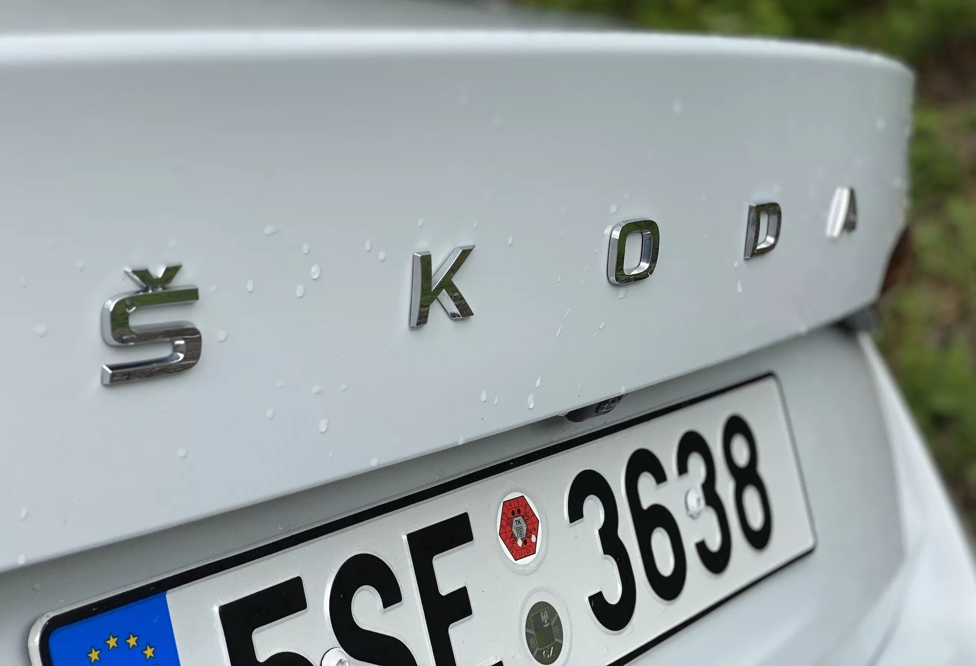 Närbild av bakre delen på en Škoda Octavia 2020 (octavia-limousine) med fokus på den kromade 'ŠKODA'-märken med en tredimensionell metallisk finish. Vinkeln på bilden är något ovanifrån och visar en svagt sluttande bagagelucka, delvis synlig europeisk registreringsskylt nedanför och regndroppar som pryder den silverfärgade bilens yta, vilket tyder på nyligen fallit regn.