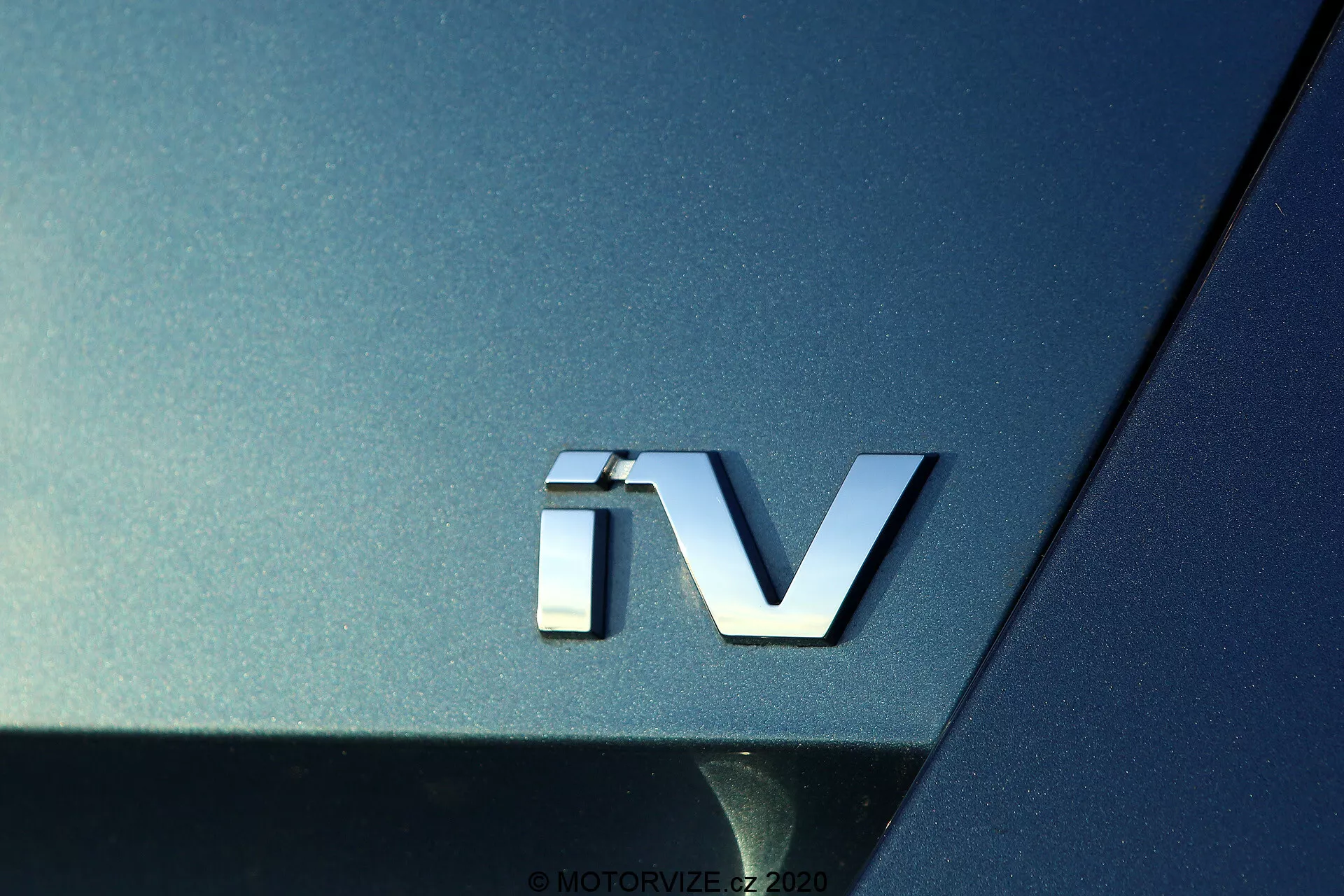 Närbild av 'iV'-emblemet på 2020 Skoda Octavia, som anger ett elektrifierat drivsystem, med ett silverfärgat 3D-emblem mot den mörka metalliska karossen, vilket framhäver den böjda ytan och de reflekterande egenskaperna hos bilens bakre sektion.