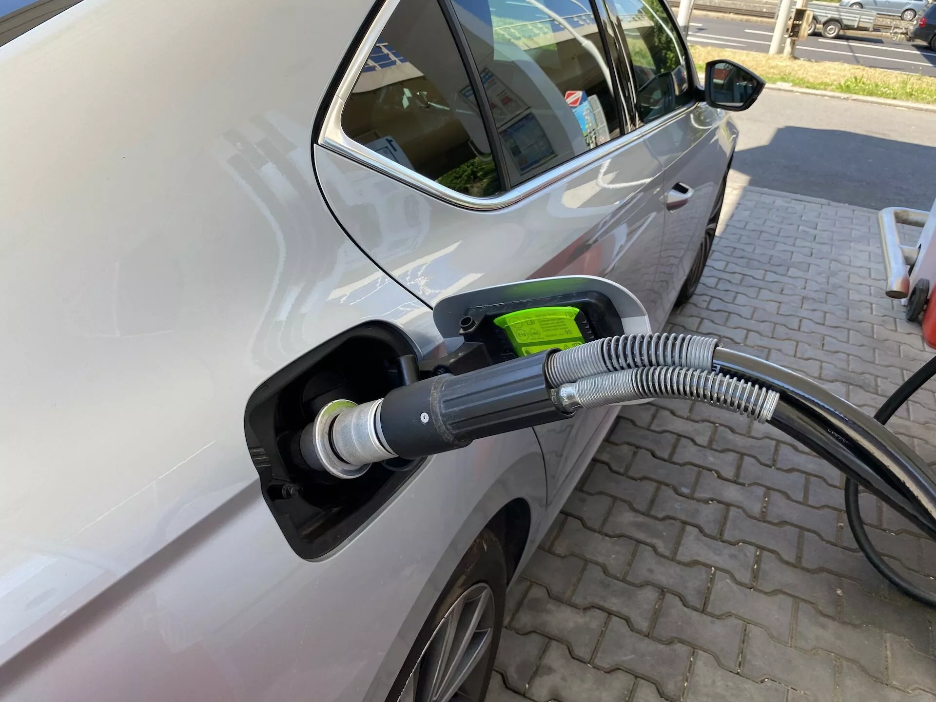 Närbild av vänster sida av en Skoda Octavia 2020 ansluten till en elektrisk laddningsstation, med fokus på laddningsportområdet och laddningskabeln och porten. Bilden är tagen från en lätt vinkel från framsidan mot baksidan, vilket visar förardörren och en del av bakdörren, vilket tyder på att det rör sig om ett fler dörrars fordon. Egenskaper inkluderar en modern sidospegel, delvis synligt lättmetallfälg, ljus bilfärg med glansigt utförande, öppen laddningsport med tjockt, korrugerat skydd på kabeln och ett grönt handtag som symboliserar miljövänlighet. Bilen är parkerad på ett belagt område med mönster av tegel, vilket antyder en dedikerad laddningsstation, utan andra fordon i närheten, vilket betonar laddningsaktionen.