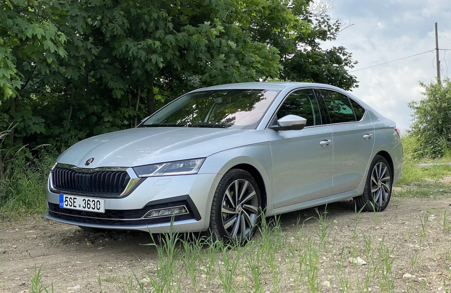 Skoda Octavia 2020 sedd från en låg främre trekvartsvinkel, med fokus på den detaljerade vänstra framsidan inklusive LED-strålkastarenheten, den framträdande grillen, främre stötfångaren med sensorer, skulpterade motorhuvslinjer, flerradiga lättmetallfälgar, integrerad blinkers i sidobackspegeln och lätt tonade fönster, placerad på en grusväg med grönt lövverk och stängsel av tråd i en lantlig eller halv-lantlig miljö.