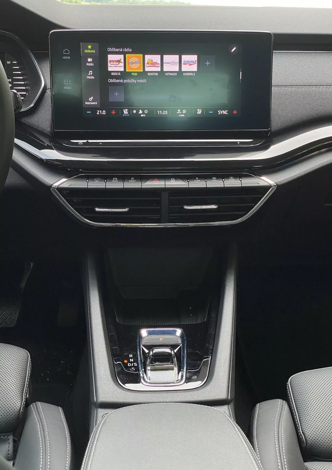 Inre perspektiv från baksätesområdet i en Skoda Octavia limousine 2020, som visar infotainmentsystemets pekskärm med färgglada ikoner för media, telefon och fordonsinställningar. Nedanför finns knappar för dubbelzons klimatkontroll med en digital temperaturdisplay, omgiven av vertikala och horisontella luftventiler. Den eleganta mittkonsolen har en blank svart växelspak med ett silverhandtag, förvaringsutrymmen med gummimattor samt synlig texturerad klädsel på framsätena i naturligt dagsljus.