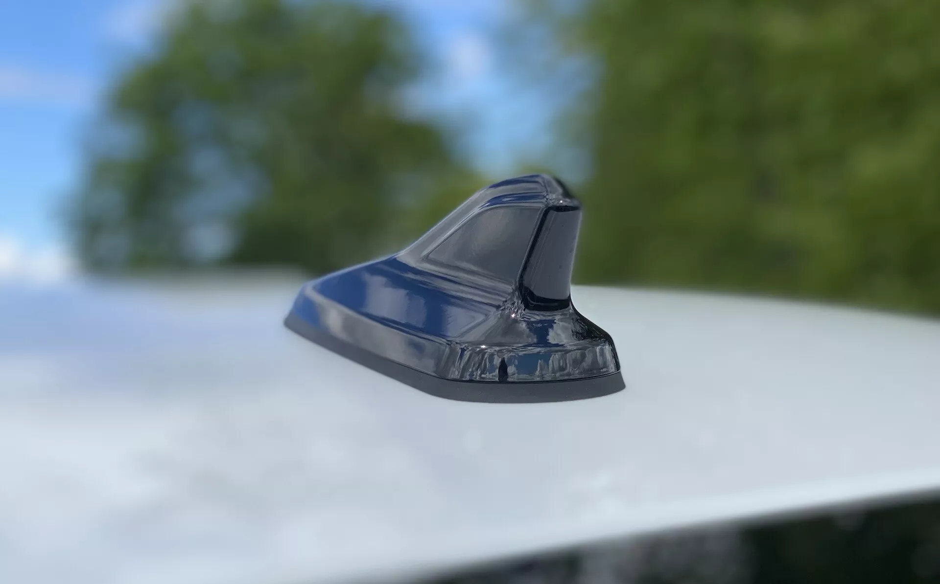 Närbild av hajfensantenn på ett fordonstak som visar modern fordonsdesign. Lågkameravinkel med kameran något lutad uppåt, fokuserar på den aerodynamiska och glänsande antennen mot en suddig bakgrund av lövverk och himmel. Den strömlinjeformade designen antyder funktioner som FM/AM-radio, satellitnavigering och avancerad kommunikationsteknik. Den metalliska lacken återspeglar fordonets väl underhållna exteriör.
