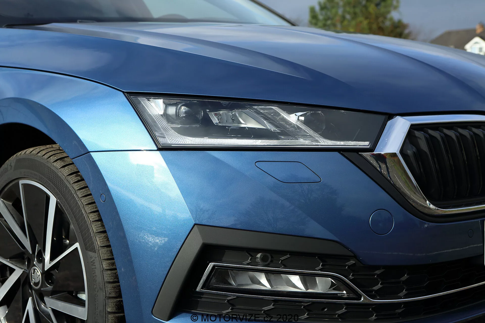 Närbild av vänster främre kvartalsdel av en 2020 Skoda Octavia limousin, som visar den moderna, strömlinjeformade LED-strålkastaren, en del av huven, vänster kant av frontgallret, den aerodynamiska främre vänstra skärmen och en del av stötfångaren med luftintag av honungskakmönstrat nät. Den metallblå lackeringen och de skarpa linjerna framhäver lyx och sportighet. Flerspokes fälg av aluminium antyder hög prestanda. Designen inkluderar en diskret märke som kan ange en specifik trim- eller teknikpaket. Denna bild fångar vackert fordonets avancerade belysningsteknik, lackfinish och prestandaorienterade designfunktioner.