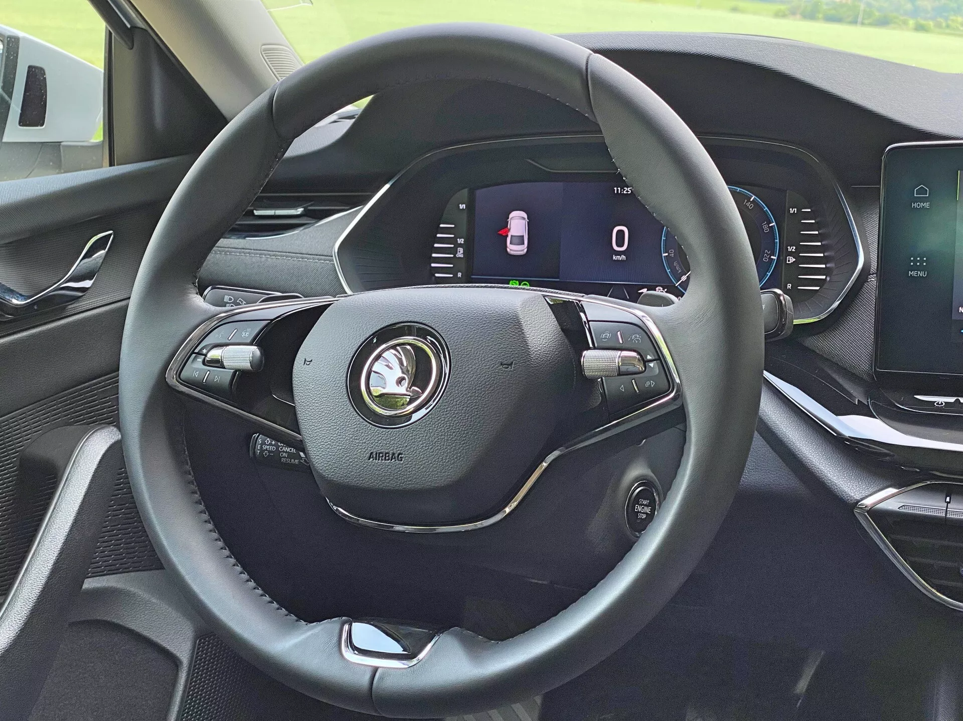 Interiören av 2020 Skoda Octavia Limousine från förarens perspektiv, med fokus på den moderna treekrade läderklädda ratten med olika kontroller för volym, röstkommandon och farthållningsinställningar. Den fullt digitala instrumentpanelen bakom ratten visar en stillastående hastighetsmätare och en indikator för öppna dörrar. Mittkonsolen har en pekskärm för infotainmentsystemet för multimedia, navigation och fordonsinställningar. Instrumentpanelen visar en modern design med högkvalitativa material.
