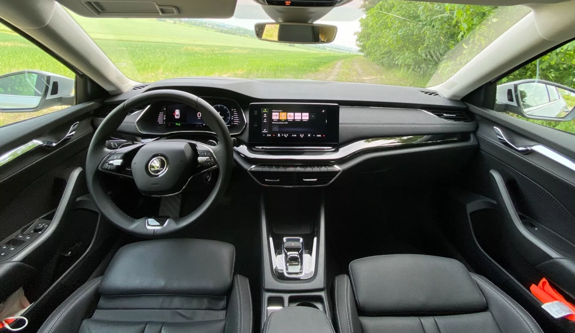 Interiörvy av en Skoda Octavia 2020 från baksätet som visar den moderna, platta bottenratten med integrerade kontroller, digital instrumentpanel, stor pekskärms infotainmentsystem på en elegant instrumentbräda, få fysiska knappar, trådlös laddningsplatta, ergonomiska framsäten i läderliknande material med kontrastsömmar och ett panoramatak som släpper in naturligt ljus i kupén.