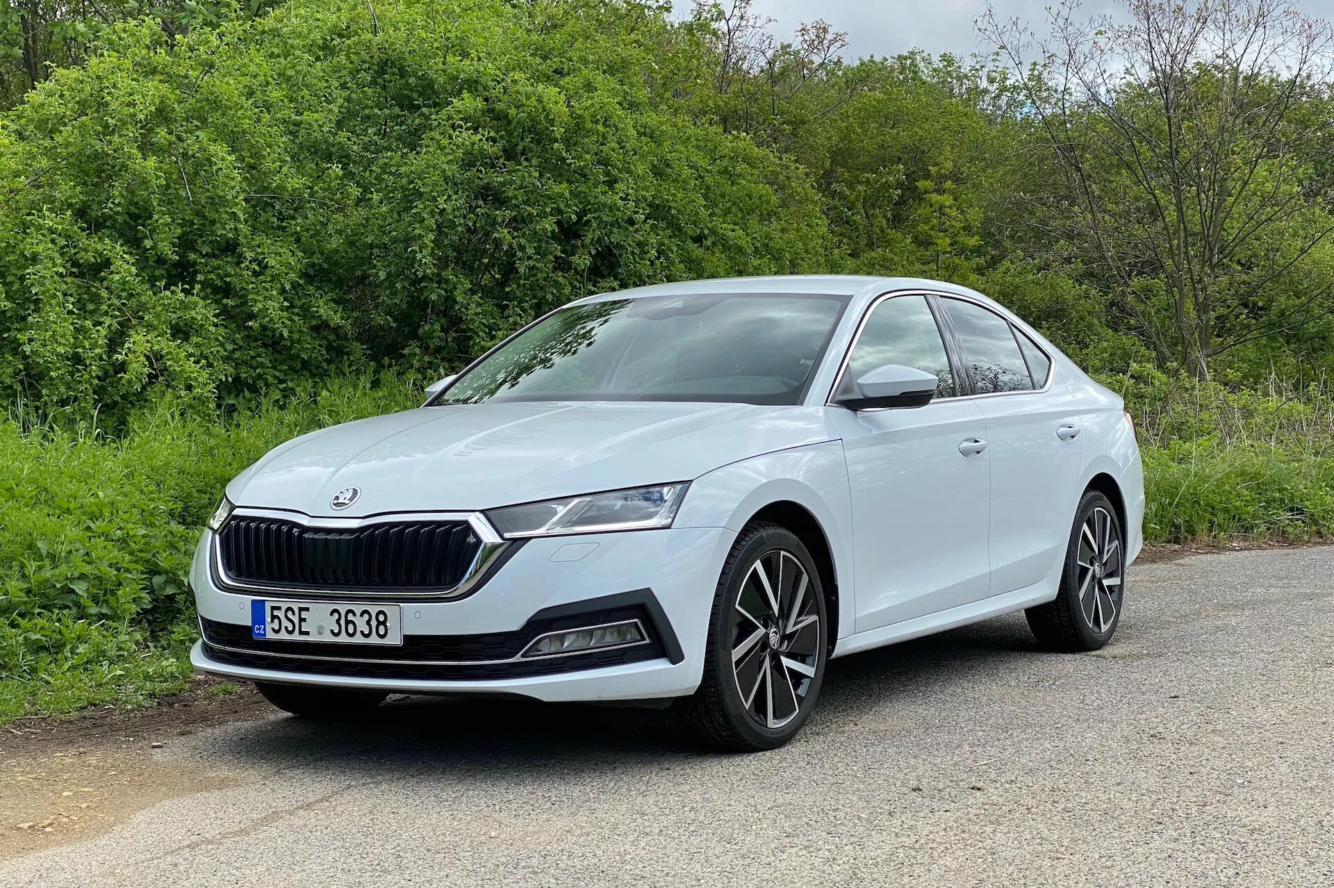 Bilden visar en Skoda Octavia 2020 i en trekvartsfrontvinkel, som visar både frontfasaden och förarsidans profil. Bilen, parkerad på en asfaltsyta med naturliga element som träd eller buskar i bakgrunden, har ett distinkt galler, eleganta LED-strålkastare, dimljushus och luftintag på stötfångaren. Fleregade lättmetallfälgar och släta, aerodynamiska sidolinjer förbättrar dess moderna och sportiga utseende. Den välbelysta och oavbrutna vyn framhäver de kroppsfärgade dörrhandtagen och den synliga karaktärslinjen längs dörrarna, vilket gör den till en utmärkt representation av bildesign.