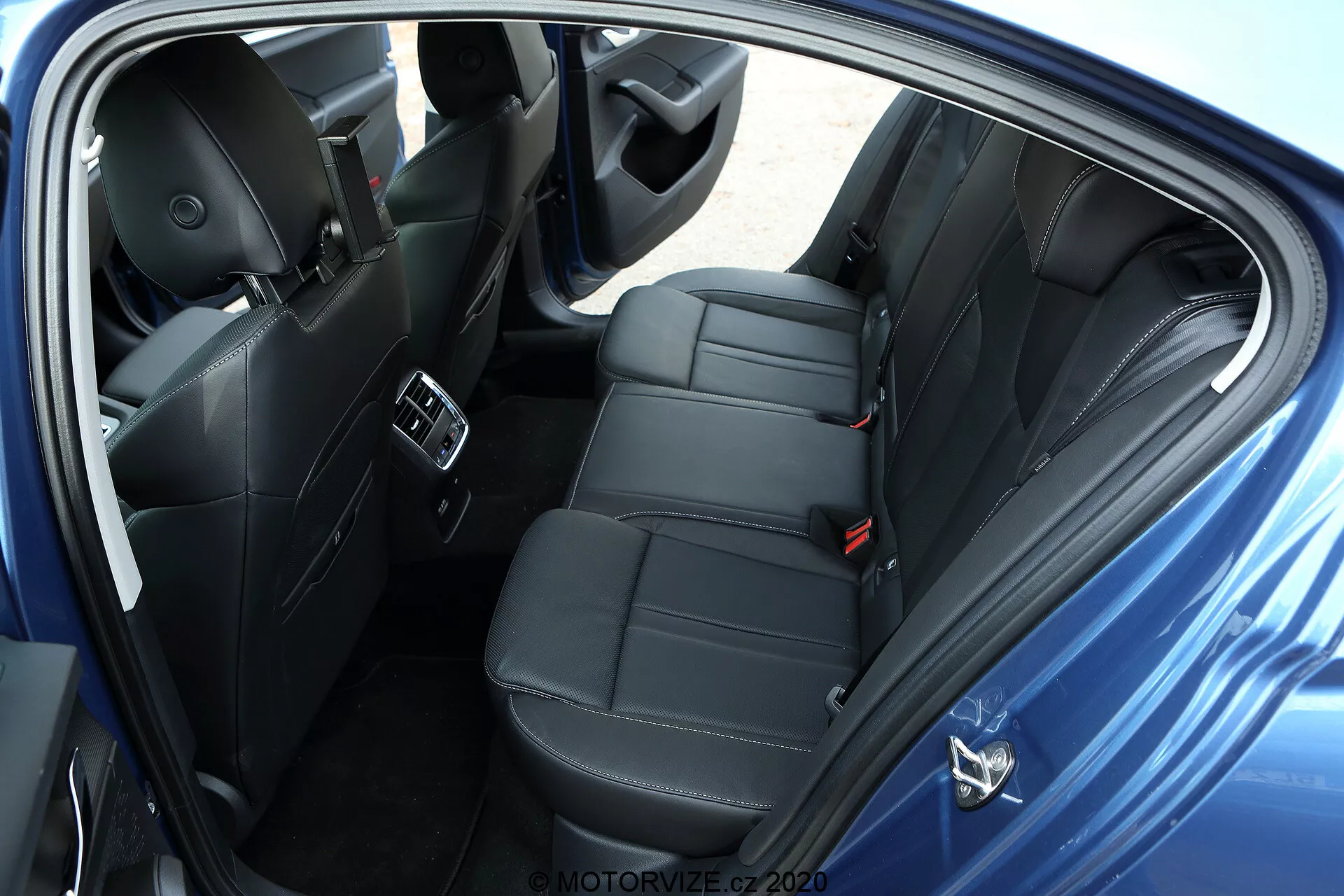 Översiktsbild över baksätespassagerarutrymmet i en Skoda Octavia 2020 limousine, tagen från höger sida. Fotot visar en öppen dörr med moderna säten i svart skinn eller skinnliknande material med lyxiga sömmar. Den bakre bänkstolen är utformad för att bekvämt rymma två passagerare med individuella nackstöd, vilket indikerar en sportigare modell eller en bekvämlighetsfokuserad trim. Bilden framhäver klimatanläggningsventiler på mittkonsolen, USB-portar för laddning av enheter, avancerade högtalare för ljudsystemet i dörrpanelen, kontroller för elektriska fönster, synliga säkerhetsbälten inklusive ett trepunktsbälte för mittsätet och mattor på golvet. Det mörka färgschemat inuti pekar på ett välutrustat premiummodellfordon utan synliga personliga föremål, parkerat med en neutral bakgrund.