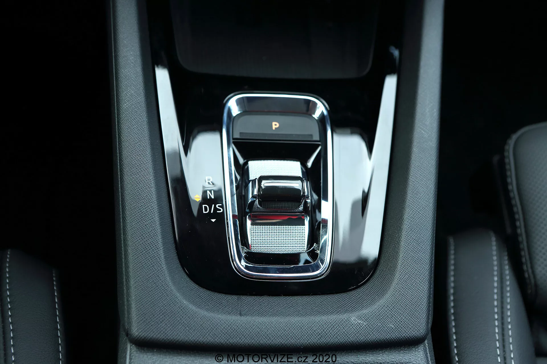 Närbild av den moderna växelspaken i mittkonsolen på en Skoda Octavia Limousine 2020. Spaken framifrån har en elegant, rektangulär design med rundade kanter och en kromkant, som visar växellägena 'P', 'N', 'D/S'. Omgiven av högkvalitativa svarta material med synliga stygn och en silverfärgad yta med fin nätstruktur, vilket antyder lyx och detaljrikedom. Ljusstark belysning lyser upp den robusta, sammanhållna interiören, vilket indikerar avancerade elektroniska funktioner och moderna bilteknologier.