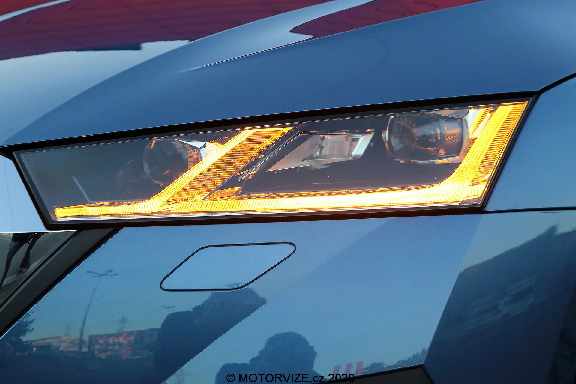 Närbild av den avancerade LED-strålkastaren på en mörkröd Skoda Octavia 2020, som framhäver dess ljusa belysning och distinkta ljusmönster. Den sofistikerade designen har ett DRL under huvudstrålkastaren, med den eleganta bilkroppen som visar skarpa kanter. Reflektioner av himlen och byggnader på den välpolerade ytan tyder på en premiumfinish. Inramningen avslöjar lätt framstöten, med fokus på innovativ bildesign.