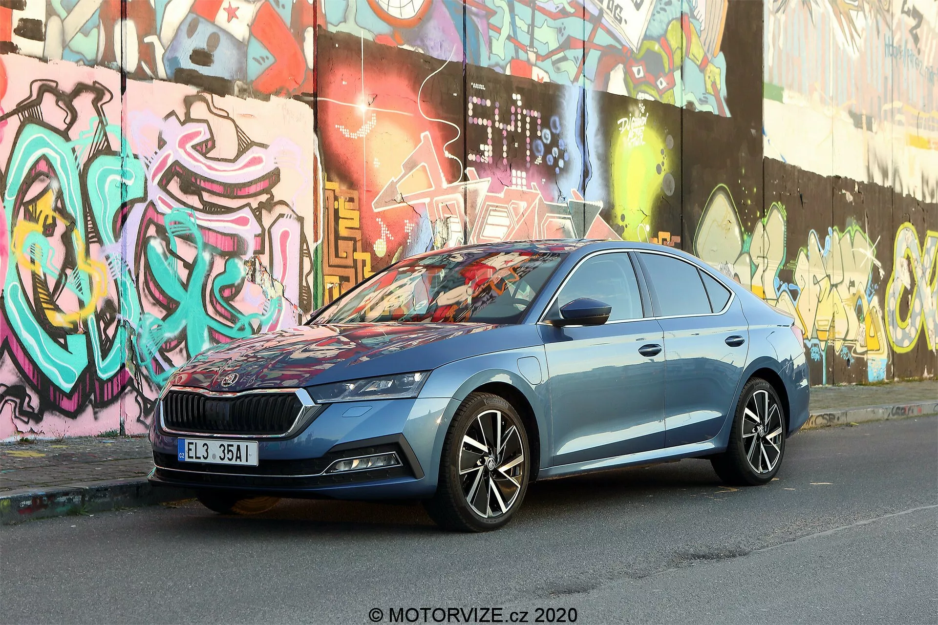 Bilden visar en 2020 Skoda Octavia Limousine parkerad på en stadsgata med graffiti-prydda väggar i bakgrunden. Fotograferad från en något låg främre trekvarts vinkel, betonar den bilens betydande grill med ett vertikalt mönster och centralt placerat emblem, eleganta LED-strålkastare, mångekrade lättmetallfälgar, dynamiska karosslinjer, integrerade blinkers i sidospeglarna, kromramade fönster och metallblå lack. Den urbana miljön framhäver fordonets moderna design och avancerade fordons-teknologi.