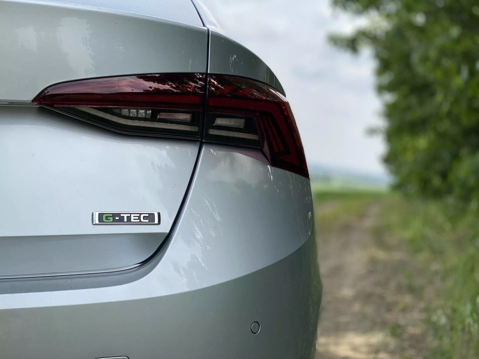 Närbild som visar den högra bakre delen av en Skoda Octavia limousine 2020. Bilden fångar designelementen på bagageluckan, LED-bakljuset och en del av den bakre stötfångaren från en sned och något förhöjd vinkel. Emblemet 'G-TEC' på bagageluckan tyder på en specifik teknologi eller utrustningsnivå. Metallfärgen framhäver bilens polerade yttre. Den suddiga bakgrunden ger en lantlig miljö som betonar bilens egenskaper och design.