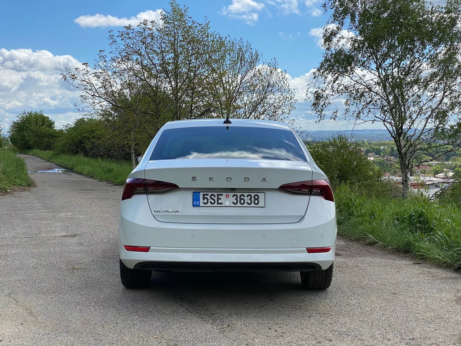 Bakvy av en vit Škoda Octavia 2020 limousine modell, stående på en smal asfalterad väg i en lantlig miljö. Bilden, tagen från marknivå i en rak vinkel, visar hela baksidan av bilen inklusive bakluckan, bakstötfångaren, bakljusen, registreringsskylten, bakrutan och Škoda-märket tydligt synligt i mitten av bakluckan med 'Octavia' på vänster sida. Runt fordonet finns träd och en molnig blå himmel, med en fjärran stad eller landskap synligt nedanför horisonten.