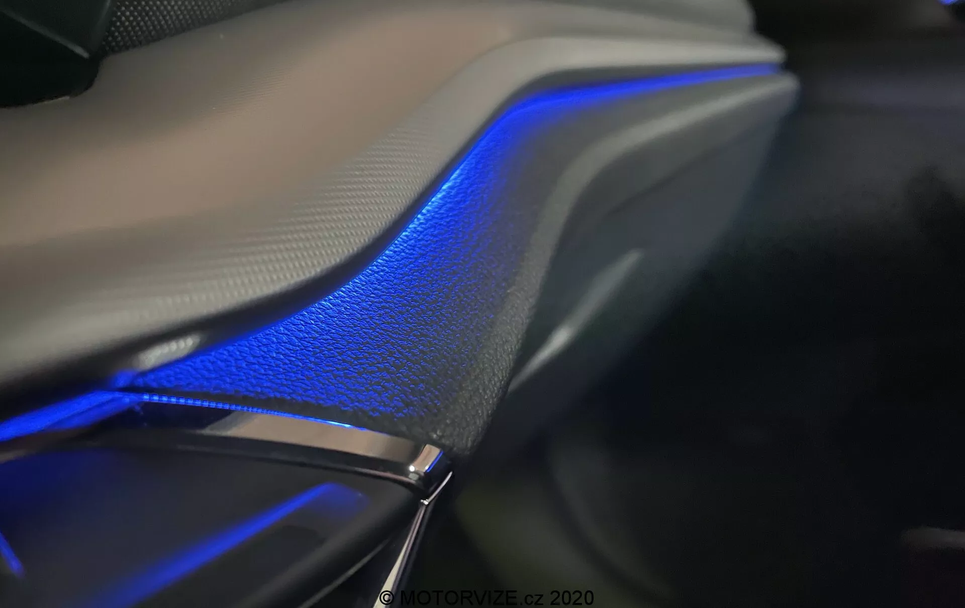 Närbild av interiören i 2020 års Skoda Octavia Limousine, som framhäver den blå ambient belysningslisten som löper längs bilens dörr eller instrumentpanel. Den eleganta designen med mjuka kurvor smälter sömlöst samman med läderytan och framhäver den lyxiga känslan under den mjuka belysningen. Fotograferad i mörk miljö, betonar bilden den blå belysningen och materialens texturer och visar upp ambient ljus teknologi som ofta styrs via bilens infotainmentsystem.
