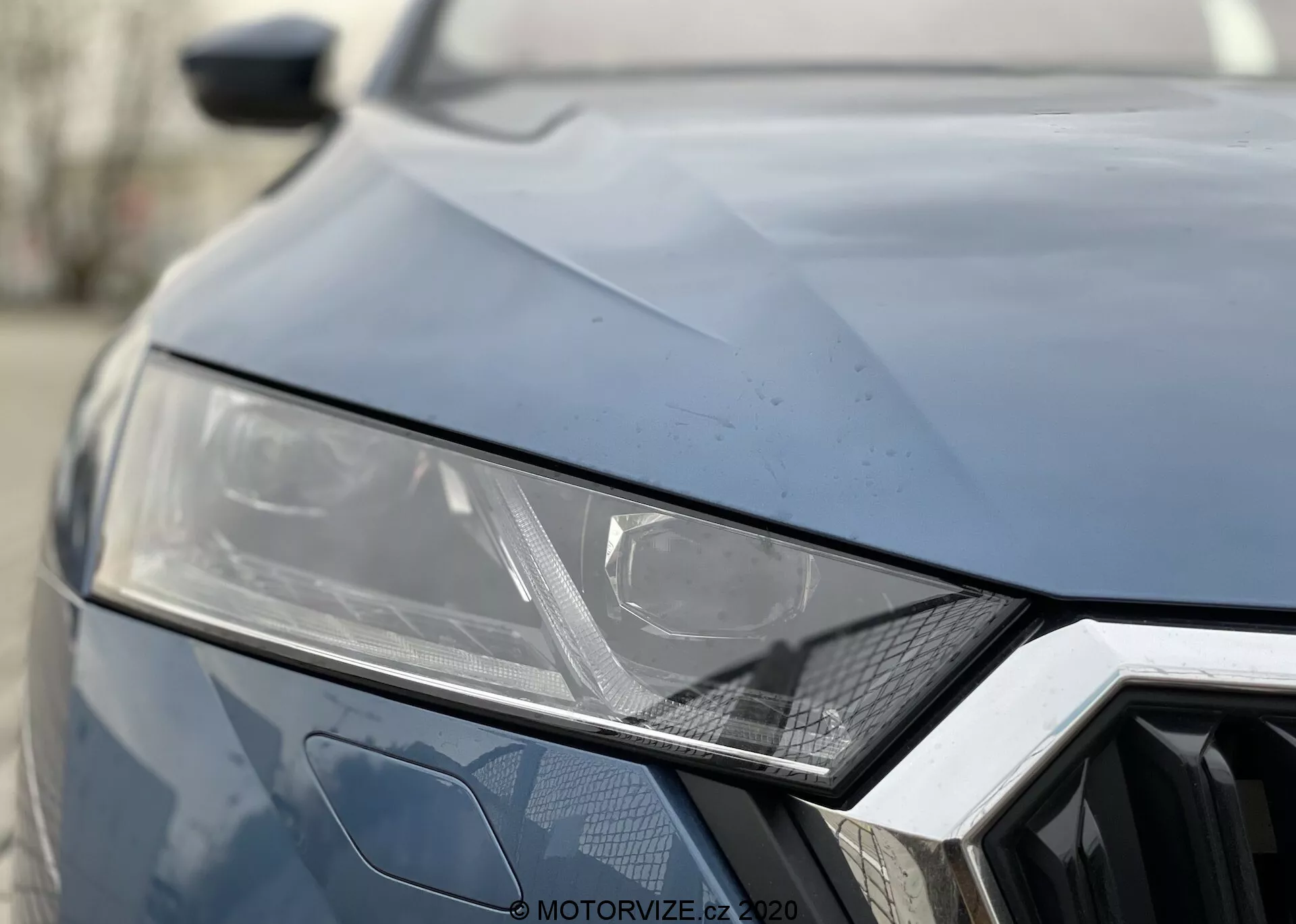 Närbild av fronten på Skoda Octavia 2020. Mörk färg, som visar vänster strålkastare med elegant, vinklad design som antyder LED- eller HID-teknologi, geometriskt hexagonmönster på grillen och vinklade veck på motorhuven för en sportig look.