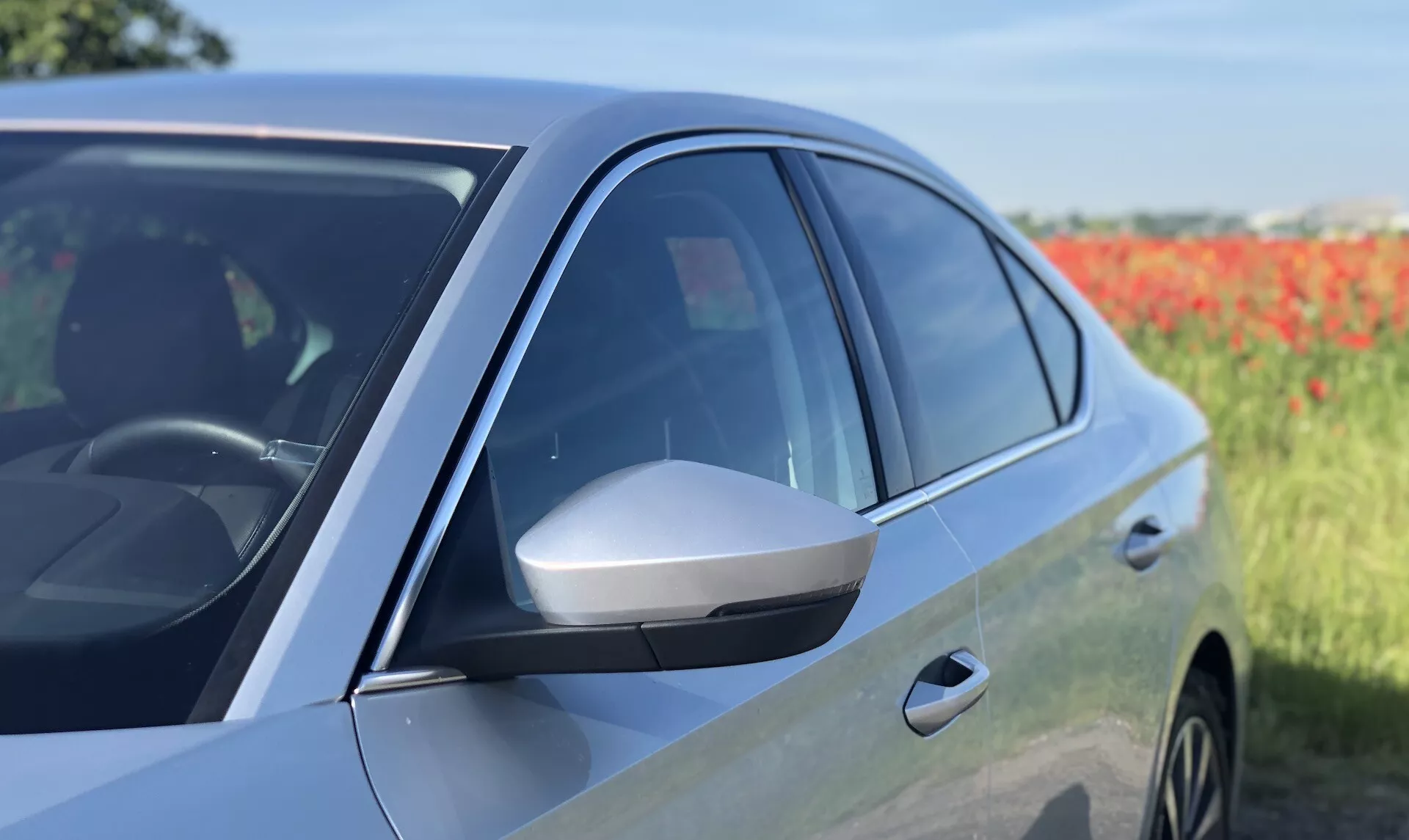 Närbild från sidan av en modern Škoda Octavia 2020 sedan, som visar förarsidans främre dörr och sidospegel. Bilden betonar den strömlinjeformade sidospegeln med integrerad blinkers, tonade fönster, glansig lackfinish och aerodynamisk design från mitten till framsidan. Reflektionen i dörrpanelen och en del av det främre hjulhuset antyder närvaron av lättmetallfälgar. Fotot framhäver avancerade designegenskaper och potentiella fordonsteknologier mot en något oskarp naturlig bakgrund.
