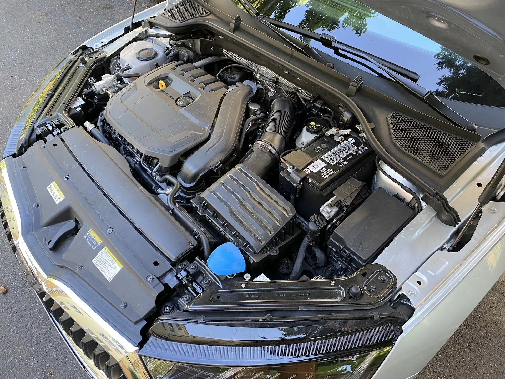 Närbild av motorutrymmet på en Skoda Octavia 2020 limousin med öppen motorhuv som visar olika komponenter inklusive den centralt inramade motoröverdraget, batteriet till höger, en rektangulär komponent med kylflänsar till vänster och en vätskebehållare med blått lock. Bilden, tagen från en fallande, något off-center vinkel, visar också den svarta plastkåpan vid basen av vindrutan, och understryker den tekniska komplexiteten i bilens motorutrymme.