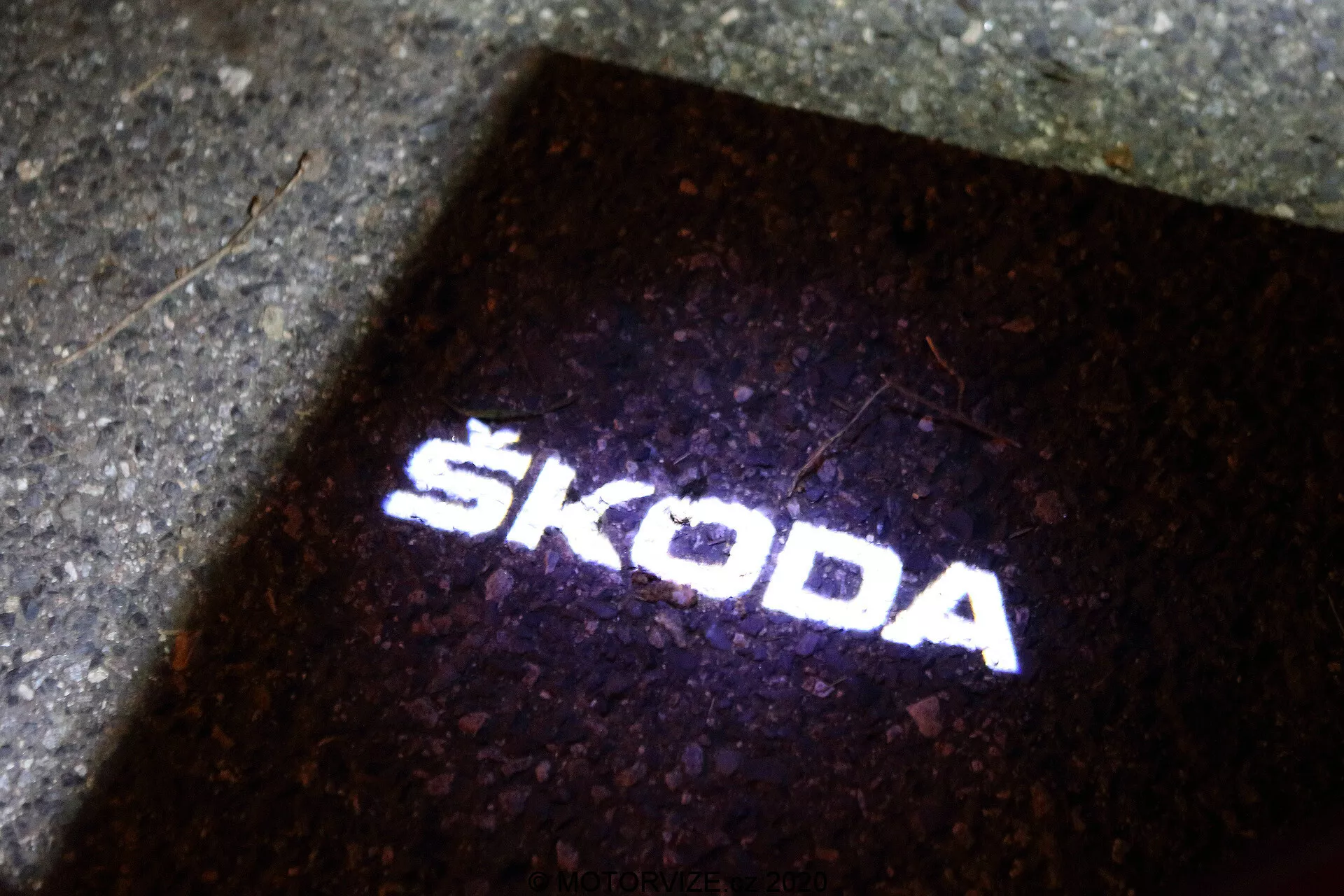 Projektion av Škoda-logotypen på marken. Ljus-källan, troligen placerad på utsidan av Škoda Octavia 2020 (octavia-limousine) bilen, skapar en 'puddle light' som lyser upp marken när fordonsdörren öppnas. Denna skarpa och tydliga vita ljusprojektion, mot en mörk bakgrund, tjänar både en funktionell och varumärkesbyggande roll, vilket indikerar närvaron av modern LED-teknik inom fordonstillbehör.