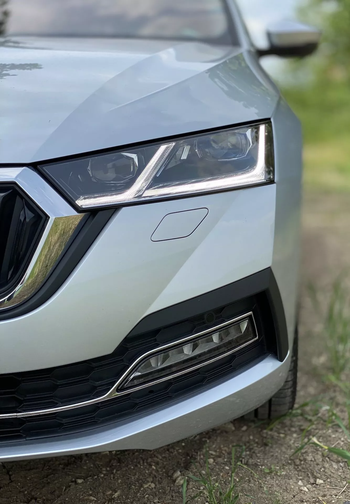 Närbild av den vänstra främre delen av Skoda Octavia 2020 (Octavia Limousine) som visar detaljer från mitten av grillen till vänster strålkastare, en del av huven och en del av den främre stötfångaren och skärmen. Bilden, tagen från en låg vinkel, framhäver den eleganta strålkastarutformningen med LED-dagljus, ramen runt grillen med kromaccenter och den nedre grillen med sin moderna metalliska finish. Fokus ligger på bilens detaljer med ett grunt skärpedjup, vilket ger en professionell, högkvalitativ utseende.