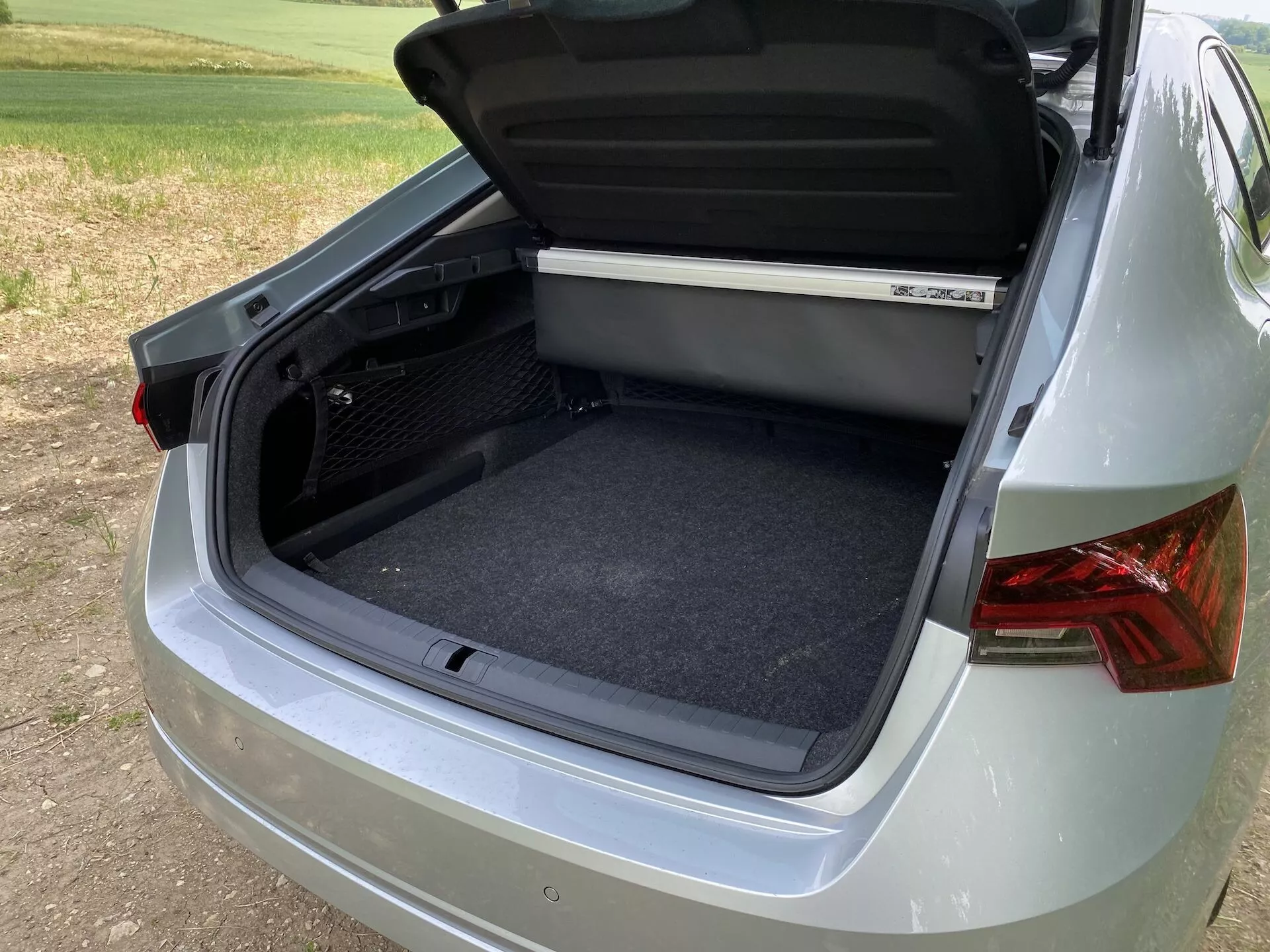 Närbild i trekvartsvy av bagageutrymmet bakom en Škoda Octavia 2020, med fokus på det öppna förvaringsutrymmet med en ren och mattbeklädd interiör. Vinkeln fångar en del av högersidan och baksidan av bilen, visar moderna bakljus, slät metalliclack och en delvis synlig stötfångare. Undersidan av bagageluckan inkluderar ett ergonomiskt handtag och en hylla syns på toppen av lastutrymmet. Miljön antyder en utomhusmiljö med mjuk dagsljusbelysning.