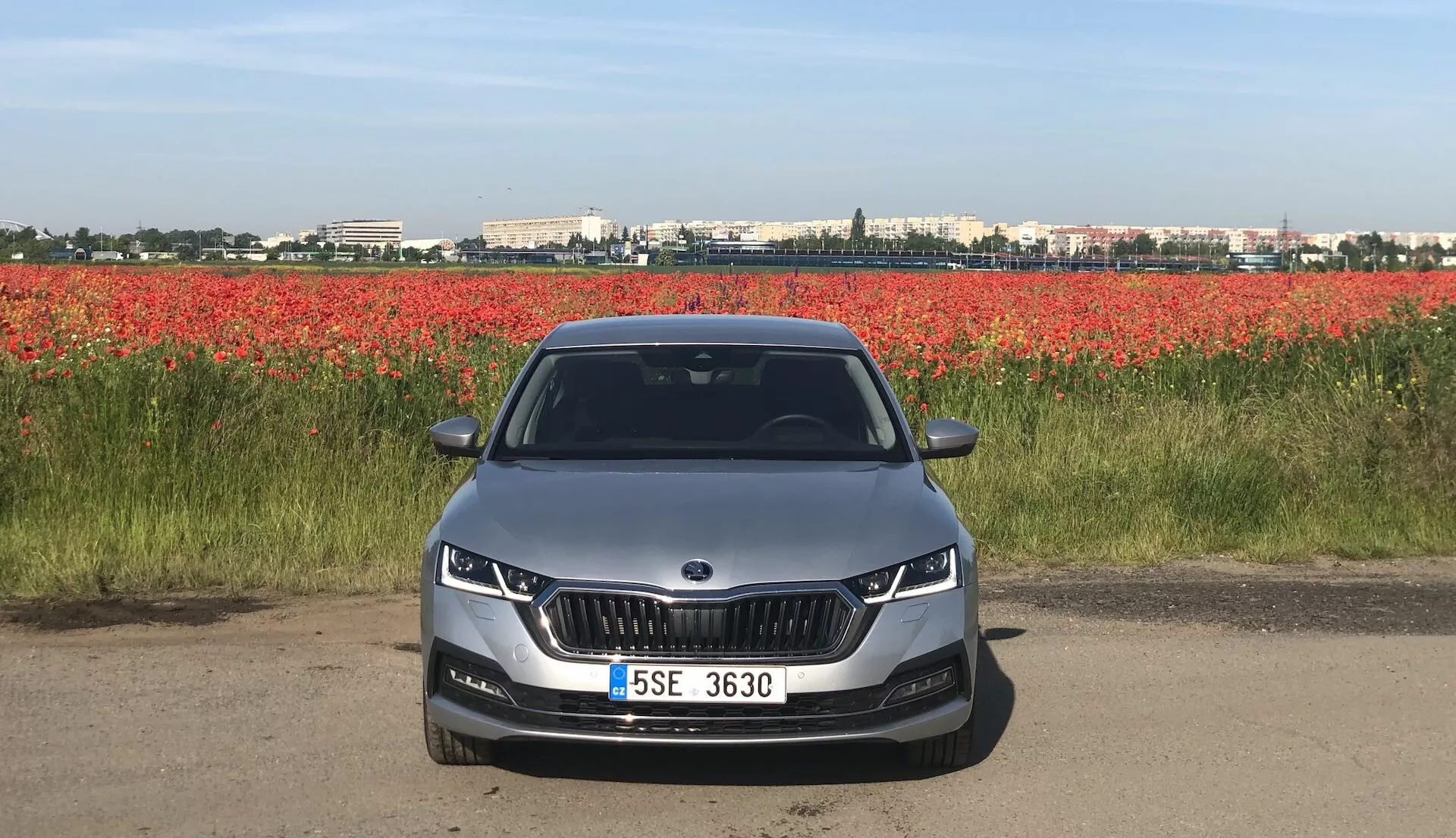Främre vy av en Skoda Octavia Limousine 2020 på en väg eller jordstig, som visar dess främre fasad med ett distinkt njurformat galler flankerat av eleganta LED- eller adaptiva strålkastare. Den aerodynamiska främre stötfångaren har utskärningar för dimljus och luftintag. Motorhuven har konturlinjer som antyder en estetik och aerodynamisk design. Den helt synliga vindrutan kan antyda avancerade teknikfunktioner som head-up-displayer eller förarassistanssystem. En ljusfärgad bil med centralt monterad registreringsskylt och parkeringssensorer, med en bakgrund av röda blommor.