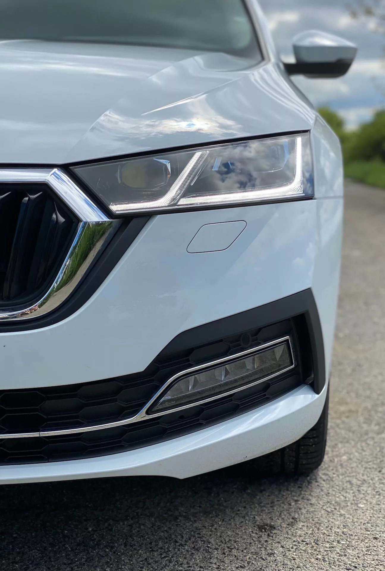 Närbild av den vänstra främre kvarten av en Skoda Octavia 2020 (octavia-limousine), som framhäver LED-strålkastaren, det distinkta grillen med kromdetaljer, den moderna främre stötfångaren med luftintag och sensorer, huvens design med veck och den delvis synliga sidospegeln med integrerade blinkers. Bilen är målad i en ljus färg, troligen vit eller silver, med smäckra karosslinjer. Bilden är tagen ur ett lågt perspektiv, fokuserad på strålkastaren och grillen, med grund skärpedjup som suddar ut omgivande element.