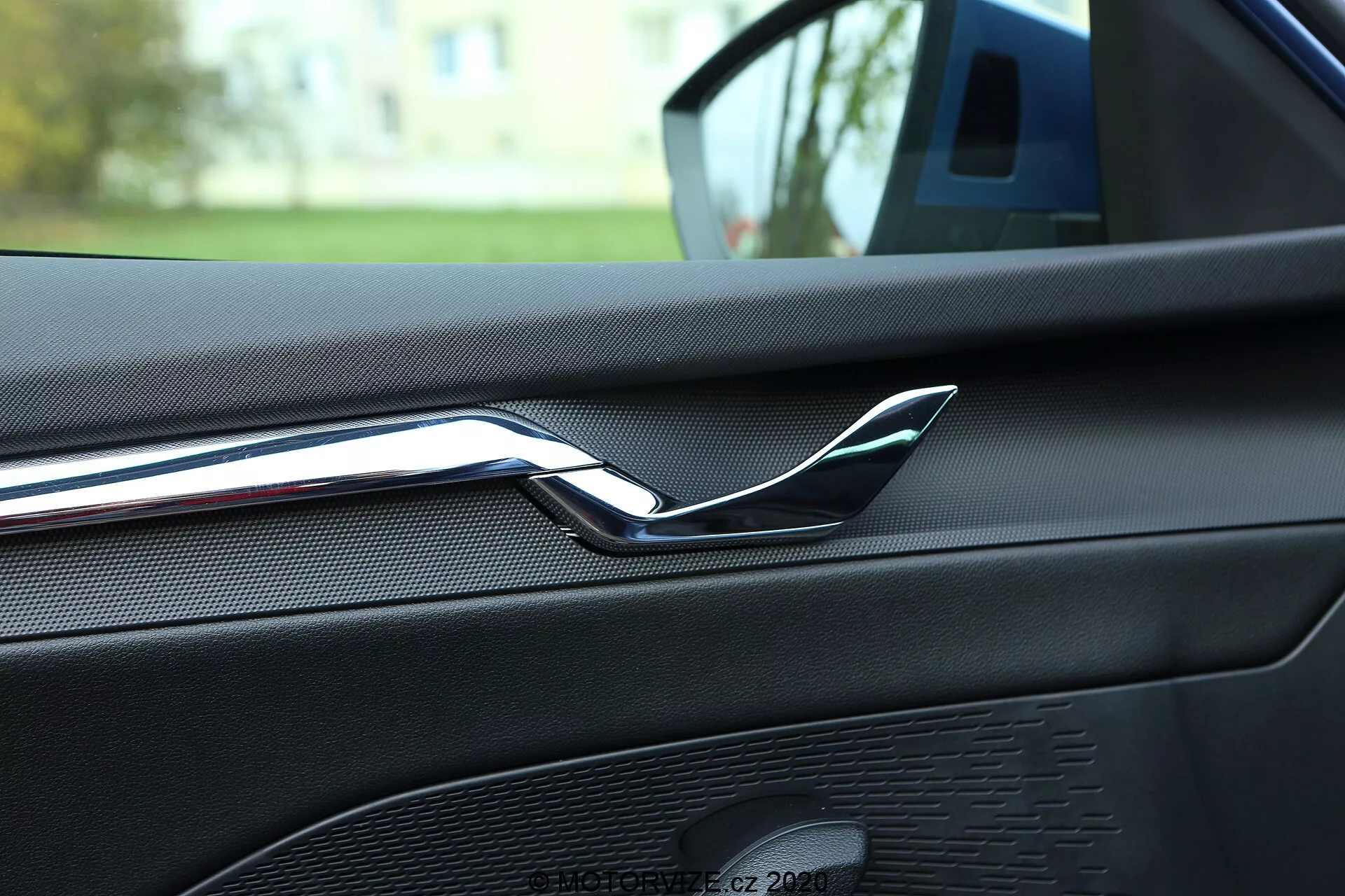 Närbild av interiören i en Skoda Octavia 2020 som fokuserar på den bakre delen av passagerardörren fram, med en reflekterande kromad dörrhandtag. Dörrpanelen är täckt med läderimitation eller texturerat mjukmaterial med synliga sömmar, vilket antyder en högkvalitativ inredning. Även synligt är ett högtalargaller som en del av bilens ljudsystem, med ett enhetligt mönster och färgkoordinerad design. Bilden är tagen från baksätet, vilket antyder att bilen är parkerad utomhus i ett bostadsområde, med grönt mjukt suddigt i bakgrunden.