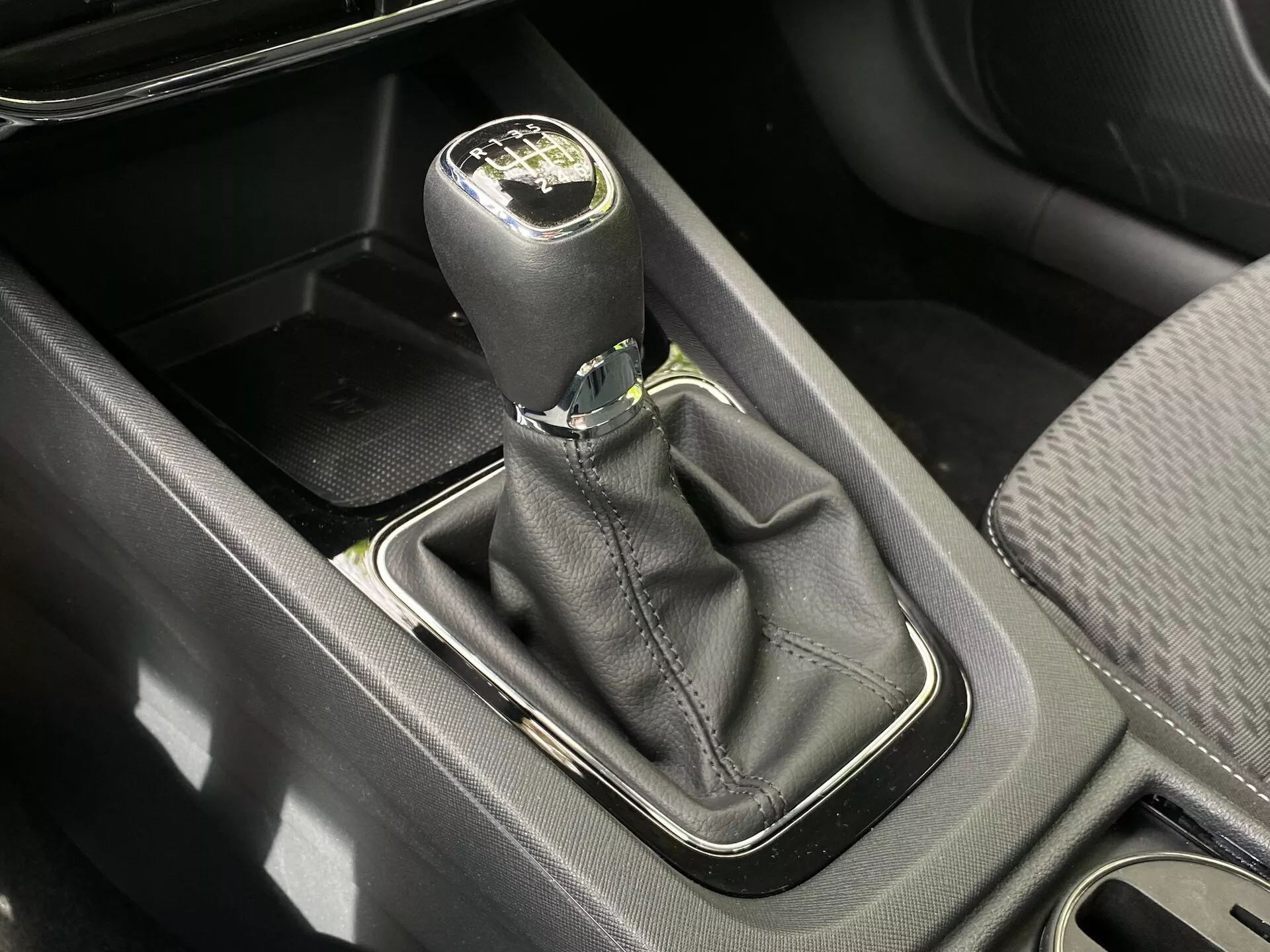 Fågelperspektiv av en manuell växelspak i en Skoda Octavia 2020 (Octavia-Limousine). Den svarta läderspaken med en silveraccent visar en layout för sexväxlad plus backväxel. Växelspakens bas, omgiven av en silverram, sitter inom en svart läderdamask. Synliga i närheten är ett förvaringsfack, mittkonsol och kopphållare, allt belyst i dagsljus.