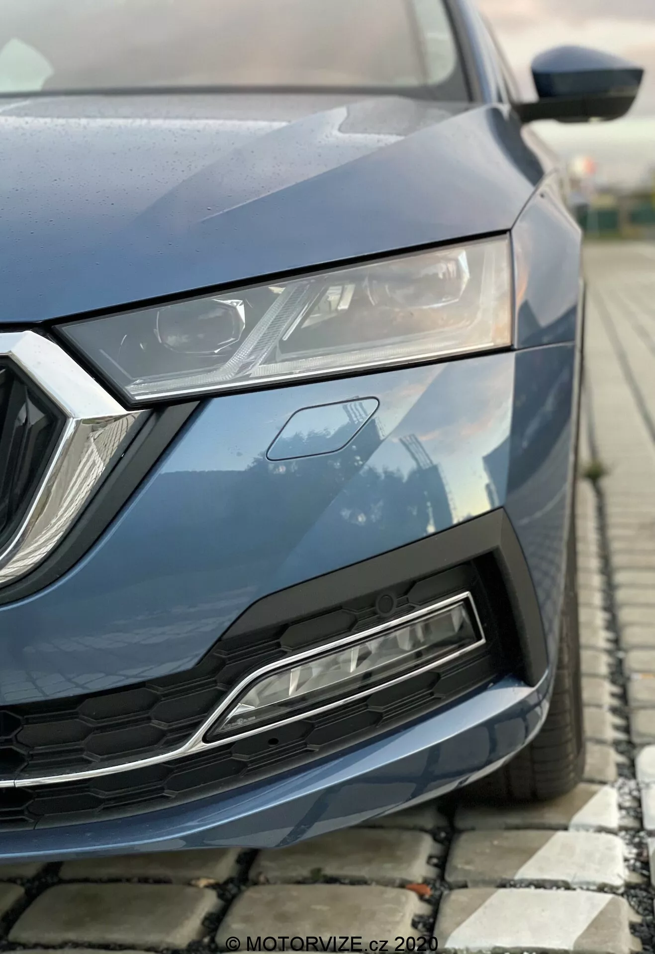 Närbild av en modern Skoda Octavia 2020 fotograferad från låg vinkel, med fokus på den främre vänstra delen inklusive motorhuv, elegant LED-strålkastare, geometriskt galler och främre stötfångare med luftintag. Metallic blå färg, kantiga designelement och framhävda aerodynamiska egenskaper.