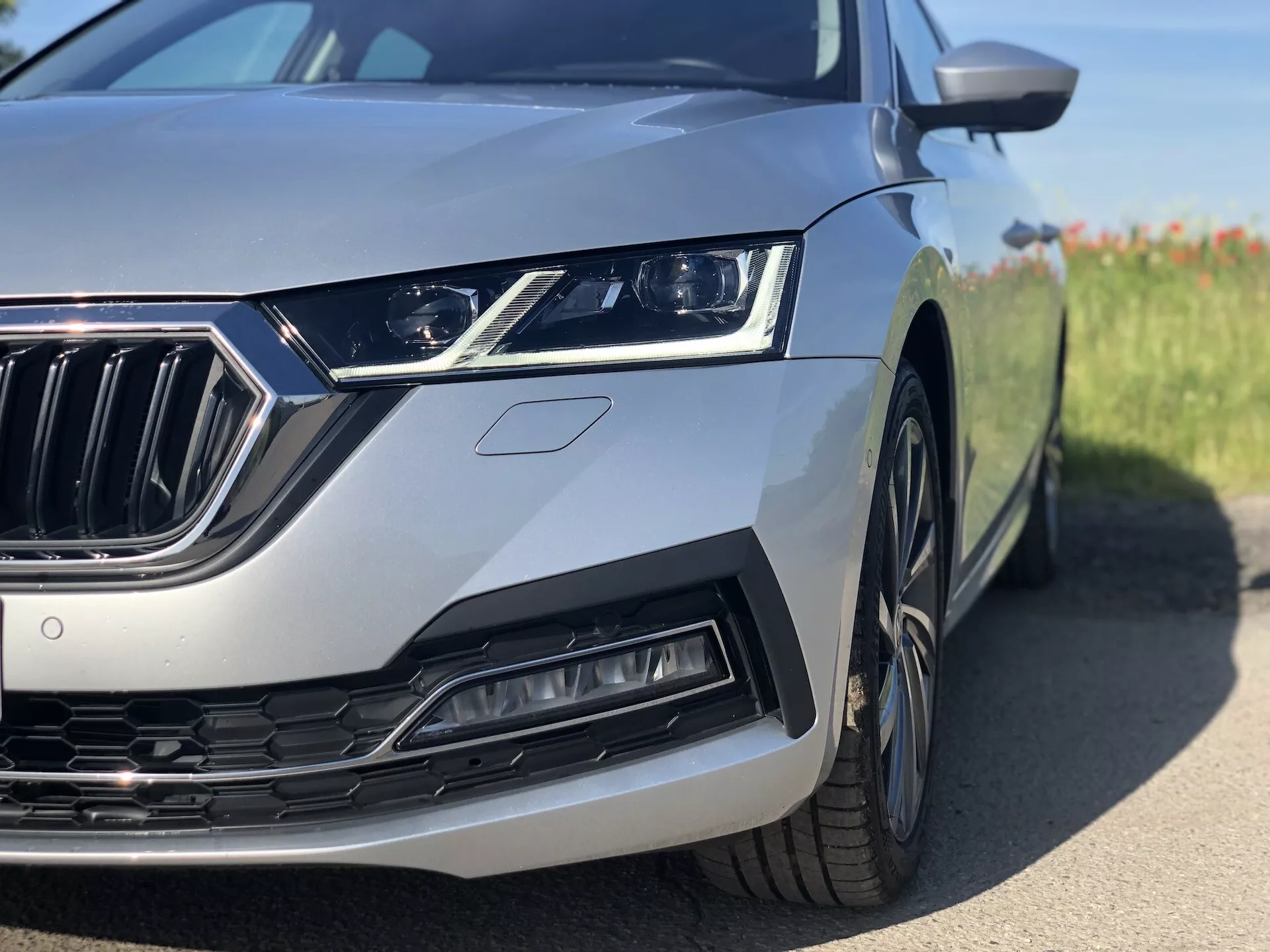 Närbild av den vänstra främre kvartdelen av en silverfärgad Skoda Octavia limousine från 2020, där den eleganta LED-strålkastaren, kromgrillen med vertikala ribbor, den skulpterade luftintaget och det delvis synliga hjulet och stänkskärmen framhävs. Bilden är tagen från en låg vinkel nära marken, med fokus på moderna designelement och avancerade tekniska funktioner mot en suddig bakgrund.