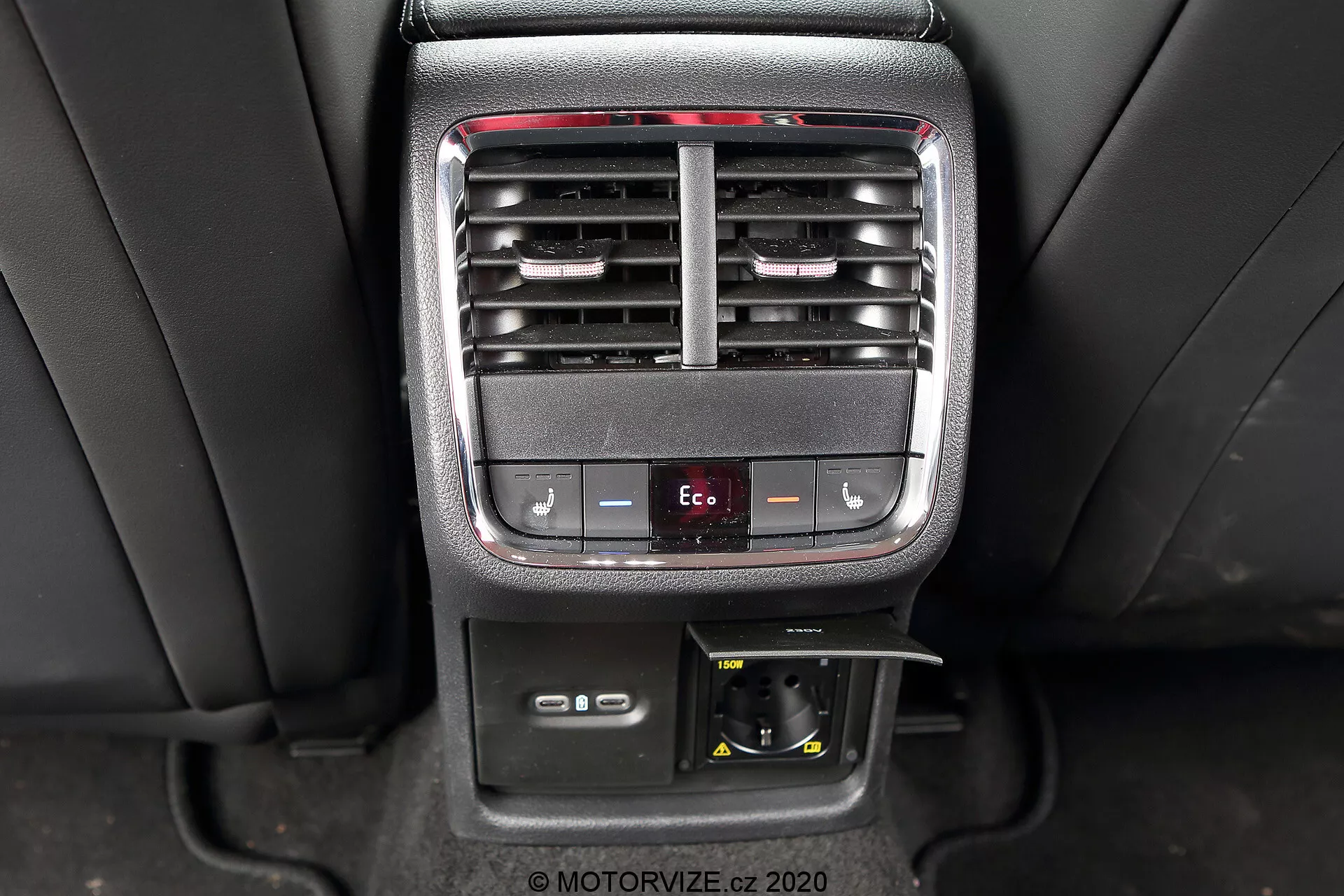 Bilden visar den inre mittkonsolen av en Skoda Octavia Limousine 2020, med fokus på bakre passagerarens klimatanläggning och bekvämlighetsfunktioner. Fotografiet ger en klar bild av justerbara ventilationsgaller, temperaturkontrollknappar, en Eco-läge knapp, ett 12V eluttag och ett förvaringsfack.
