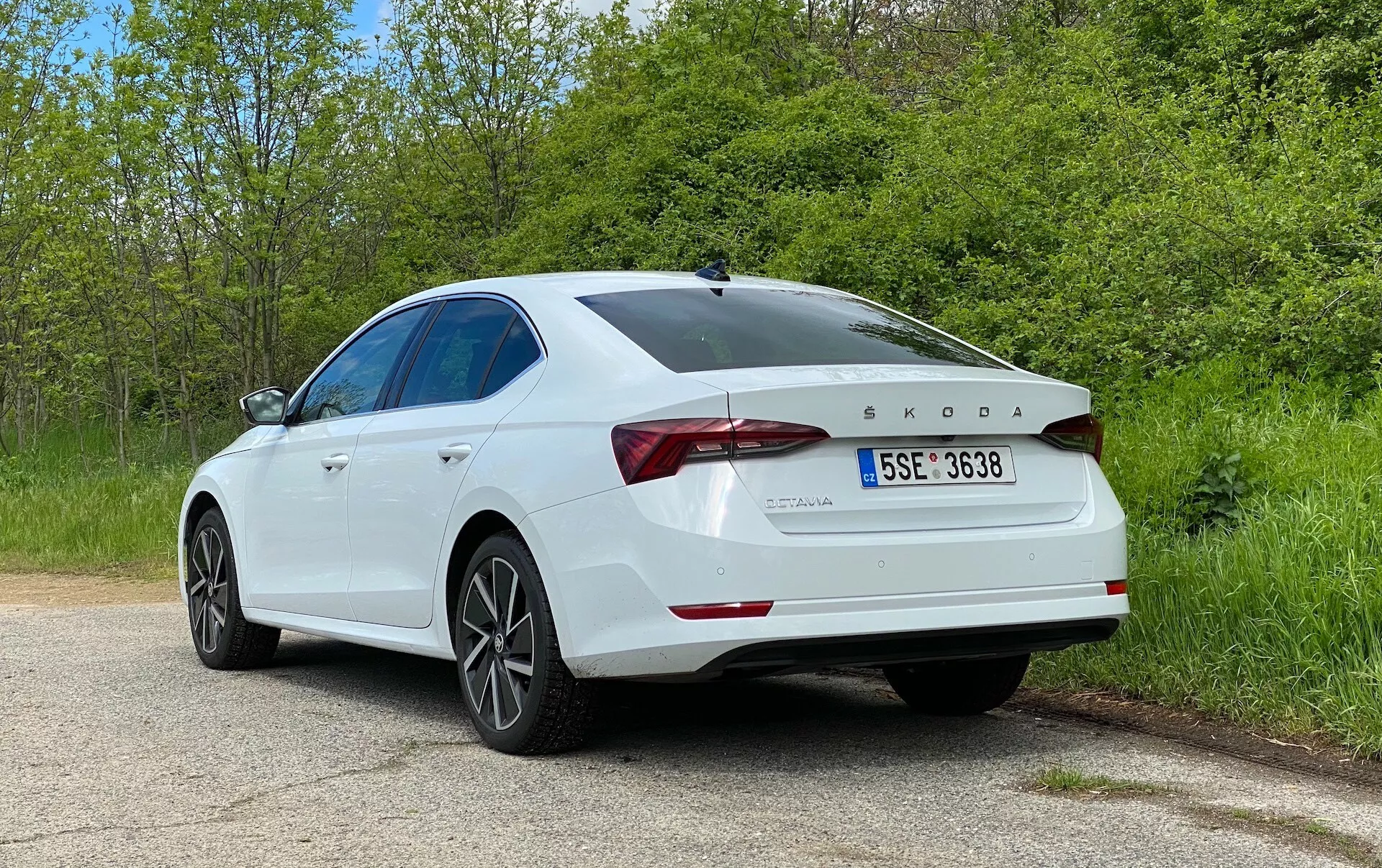 Bilden visar en vit Škoda Octavia 2020 sedan tagen från en trekvartsvinkel bakifrån, vilket framhäver fordonets högra sida och bakre komponenter. Nyckeldetaljer inkluderar LED-bakljus, Škoda-logotypen och OCTAVIA-modellens emblem, TDI-märkning, skulpterat bagagelucka med integrerad spoiler, femekrade lättmetallfälgar, slät sluttande taklinje, bakre stötfångare med parkeringssensorer och glansig, väl underhållen lack. Bilen är parkerad på en lugn landsväg eller parkväg med grönska på båda sidor, vilket förstärker den fridfulla miljön. Detta foto betonar bilens yttre design, moderna sedanfunktioner och tekniska element som dieselmotorn och förarassistanssystemen.