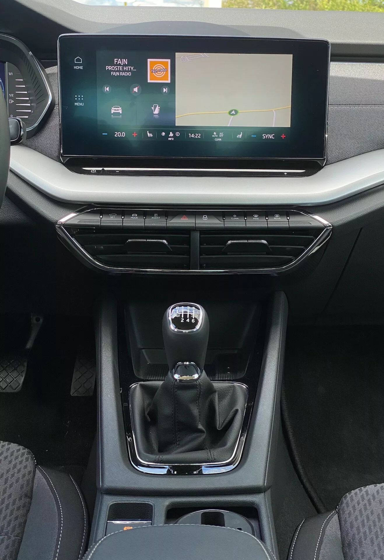 Detaljerad vy av det centrala inre konsolområdet i en Skoda Octavia limousine 2020 från baksätets perspektiv, som visar en bred infotainment pekskärm med medie- och klimatkontroller, ventilöppningar med metallaccent och minimalistiska designknappar, traditionell manuell stil automatisk växelspak med tecken för manuellt läge, förvaringsutrymme med USB-portar, mugghållare och en blandning av läder och släta plastfärdiga ytor som betonar modern komfort och förarcentrerad användbarhet.