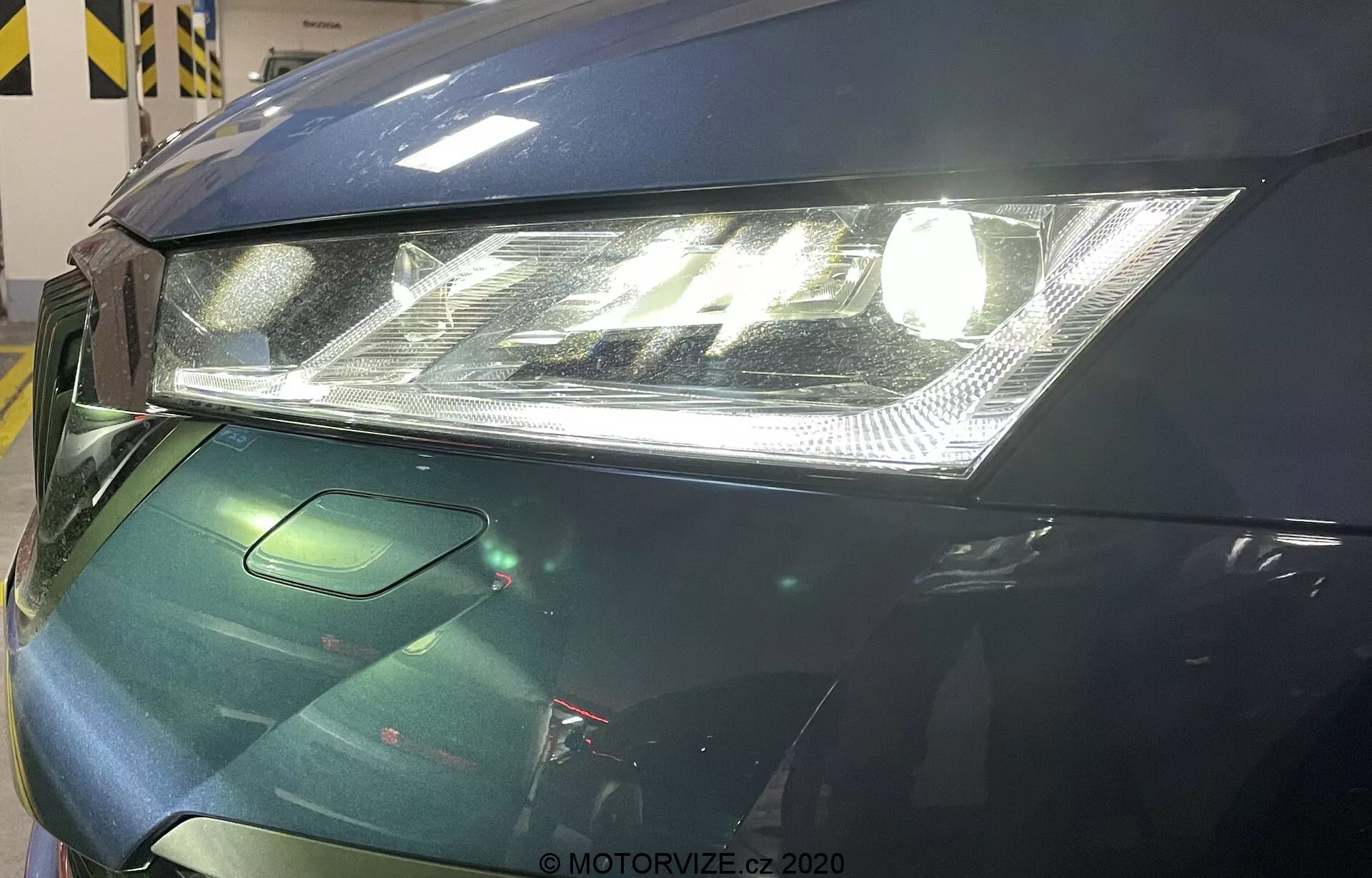 Närbild av den främre vänstra delen av en Skoda Octavia 2020. Bilden fokuserar på en modern strålkastare med klar linsteknik och integrerade LED-dagljus, en delvis synlig huva med smidig design och en liten del av grillen som troligen innehåller sensorer för förarehjälpssystem. Bilen har en mörkgrön, glansig finish som antyder avancerade lackskyddsåtgärder.
