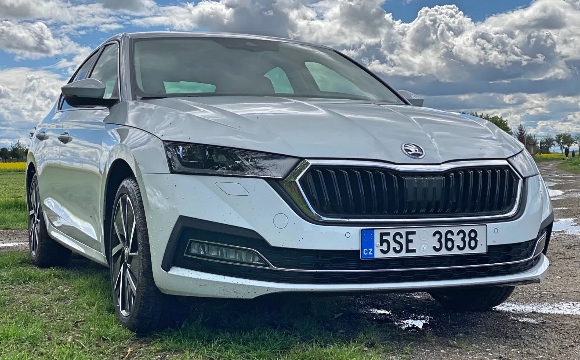 Låg vinkel framifrån på en vit Škoda Octavia 2020 parkerad utomhus, med fokus på fronten, skarpa strålkastare med LED-dagsljus, bred grill med Škoda-logga, parkeringssensor, sportigt främre stötfångare med dimljus, fler-ekrade lättmetallfälgar och integrerad blinkers i sidospegeln. Den panoramiska vindrutan och den europeiska registreringsskylten är också synliga.