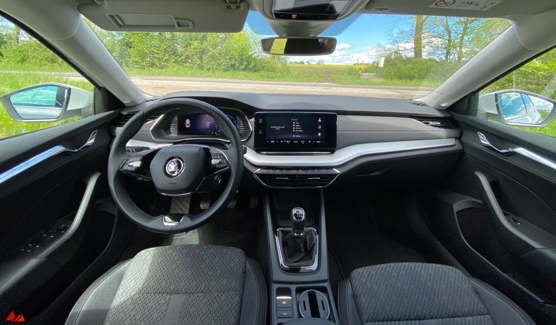 Interiörvy av Skoda Octavia 2020 från förarens perspektiv, som visar den multifunktionella ratten, digitala instrumentpanelen, horisontellt orienterad infotAINMENT-pekskärm, minimalistisk mittkonsol med växelväljare, mugghållare och mjukt beröringspanel med träliknande trim under ljusa dagsljusförhållanden.