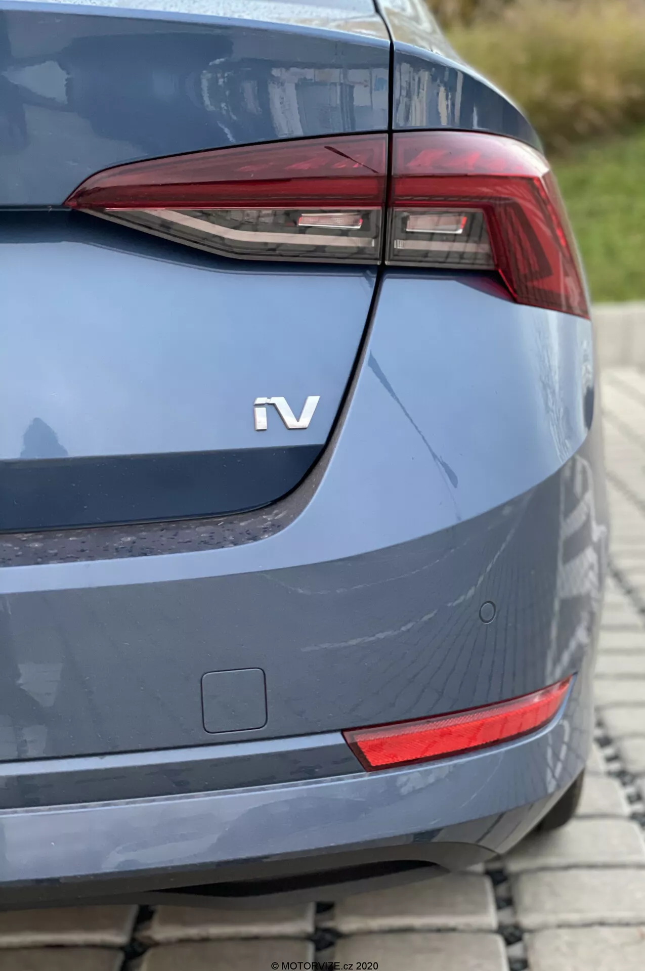 Närbild av bakdelen på en Skoda Octavia 2020 från en sned vinkel, med fokus på det moderna, kantiga bakljuset troligen med LED-element. Under bakljuset syns en liten rektangulär reflektor eller extra ljusenhet. Den metalliska blågrå karossen har ett milt böjt bagagelucklock, välintegrerad bakre stötfångare och en 'iV'-märke som indikerar en hybrid- eller eldrivlina. Nästan osynliga parkeringssensorer i det nedersta högra hörnet tyder på avancerade förarassistanssystem. Fotot, taget i dagsljus med ett kort skärpedjup, visar bilen parkerad på en kullerstensyta.