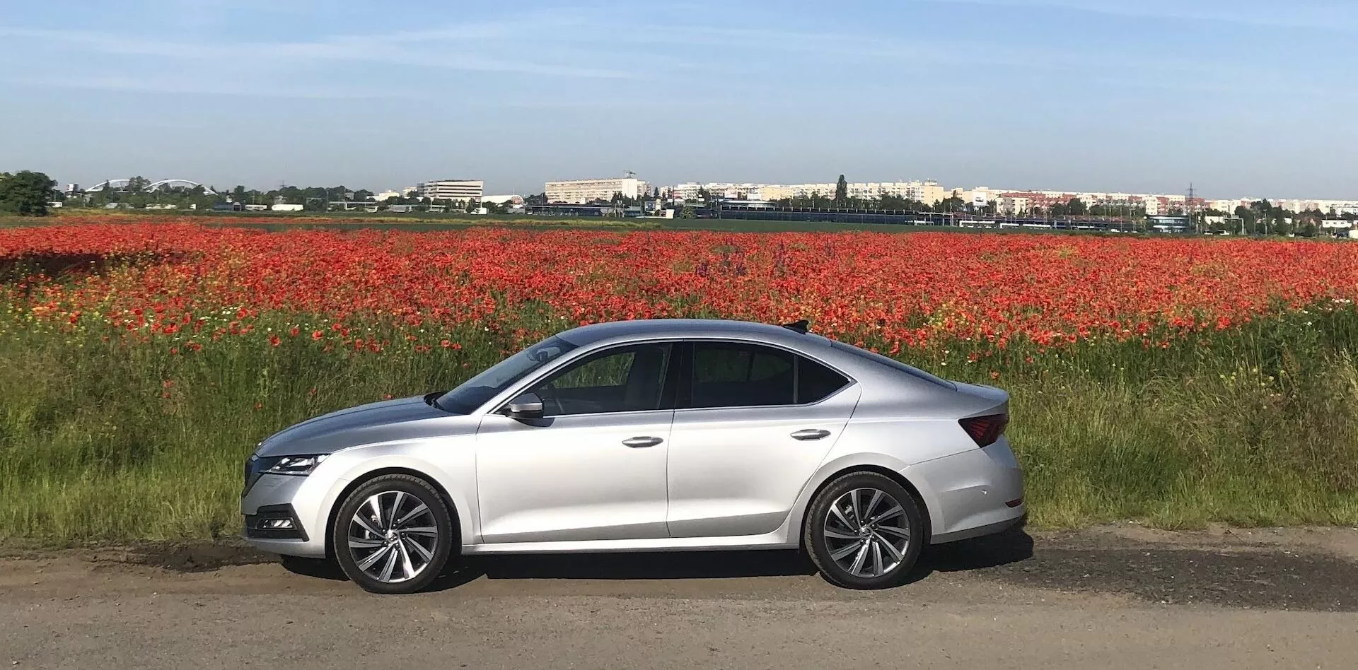 Sido vy av en modern silverfärgad Skoda Octavia 2020 sedan parkerad på en plan yta bredvid ett fält med röda blommor, med elegant aerodynamisk design, LED-strålkastare, karossfärgade sidospeglar med integrerade blinkers, lättmetallfälgar, fyra dörrar, tonade rutor och en mjukt sluttande taklinje.