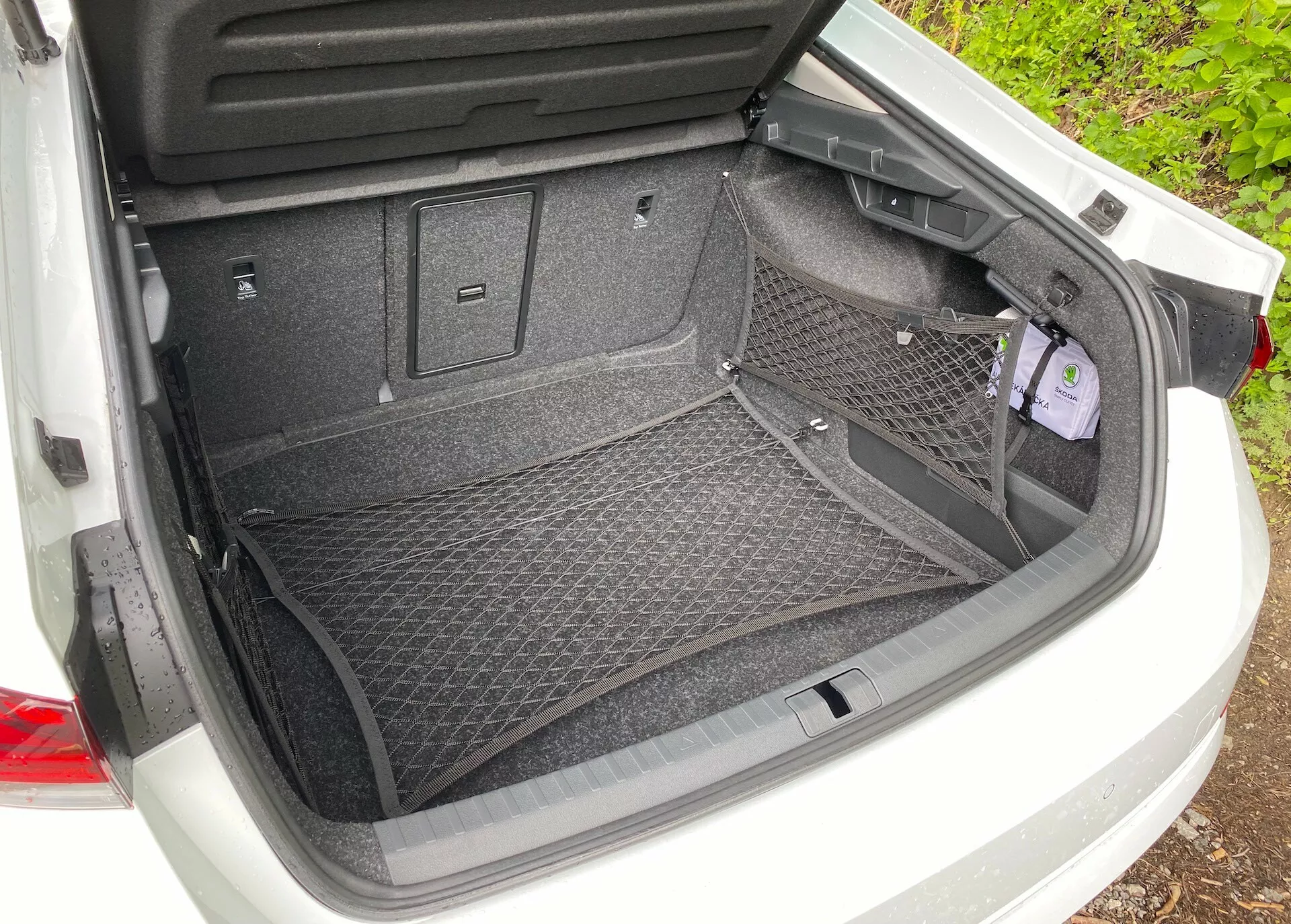 Bakifrån syn av det öppna bagageutrymmet i en Škoda Octavia 2020 (octavia-limousine) med en mattbelagd yta och lastnätstillbehör för säker förvaring. Funktioner inkluderar plastklädsel för skydd, olika krokar och fästpunkter för extra lastalternativ. Till höger finns ett litet nedsänkt fack med lock som indikerar extra förvaring eller tillgång till viktiga verktyg.