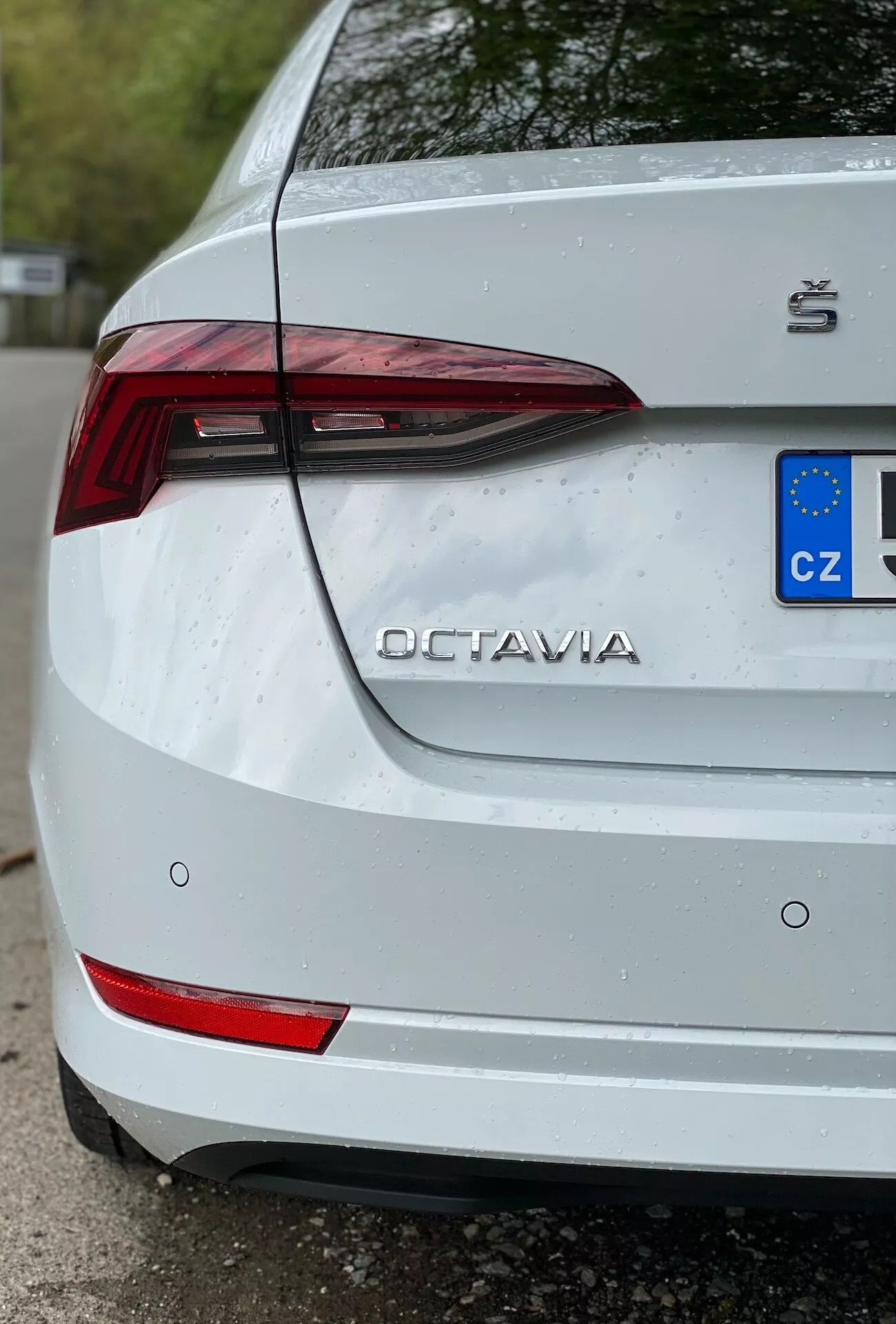Detaljerad närbild i trekvartsperspektiv av bakdelen på en vit Škoda Octavia 2020 (limousine), med fokus på höger sida. Bilden framhäver framstående detaljer som den vinklade LED-baklyktan, reflexelement på den bakre stötfångaren, 'OCTAVIA'-namnet och Škoda-logotypen på bagageluckan samt delvis synlig EU-registreringsskylt med 'CZ'-beteckning. Tunna vattendroppar på ytan antyder nylig exponering för regn eller tvätt, vilket förbättrar fräschheten hos den vita färgen medan den visar lätt smuts kring registreringsskylten.