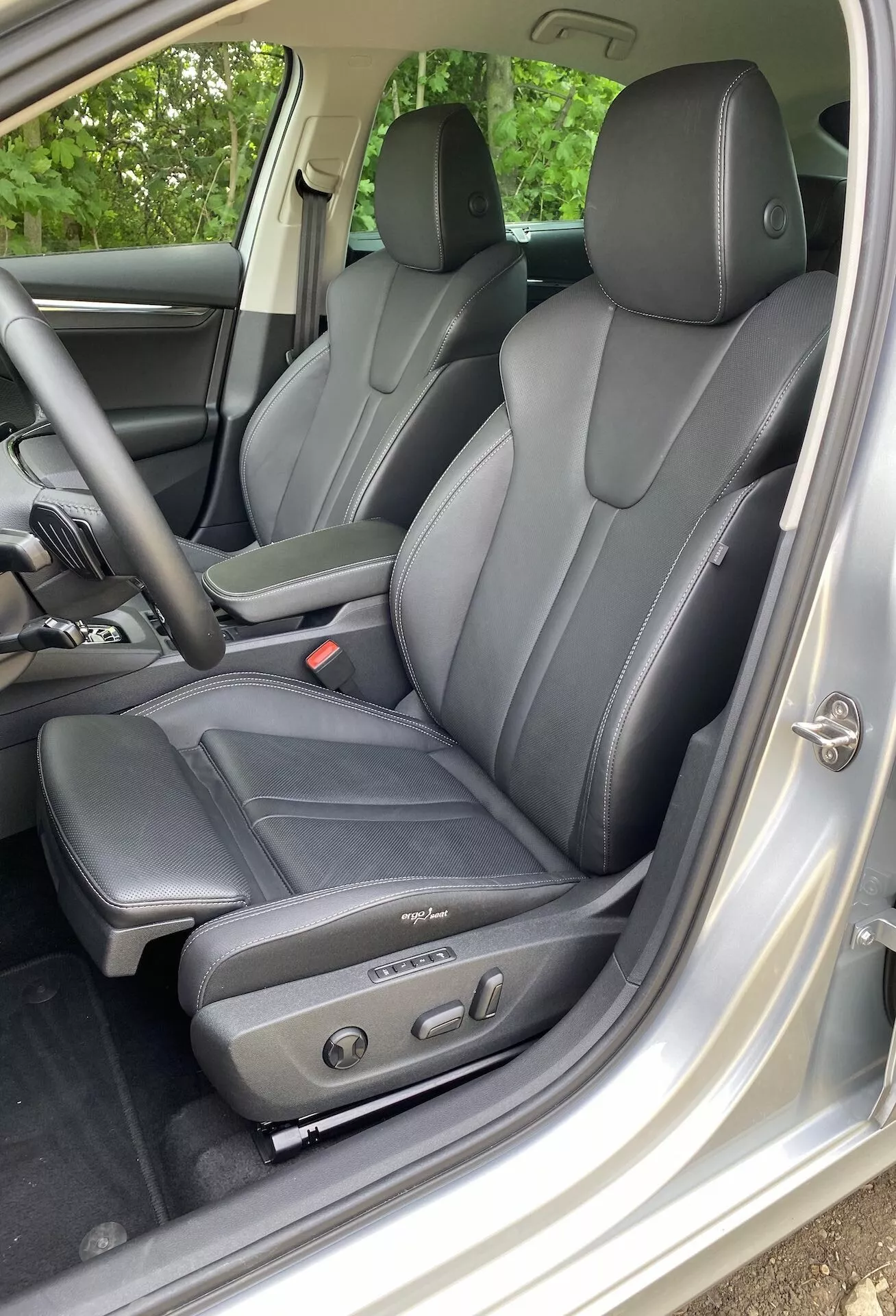 Interiörvy av 2020 Škoda Octavia limousine fokuserad på förarområdet, med mörka lädersäten med ergonomisk design och elektriska justeringskontroller, sedd från vänster sida genom den öppna förardörren. Synliga nackstöd för förare och baksätespassagerare indikerar plats för minst fyra passagerare. Den inre trimmen i silver eller ljusgrå kontrasterar med sätena och dörrpanelen inkluderar kontroller för eldrivna fönster och speglar, vilket betonar teknologisk komfort och lyx.