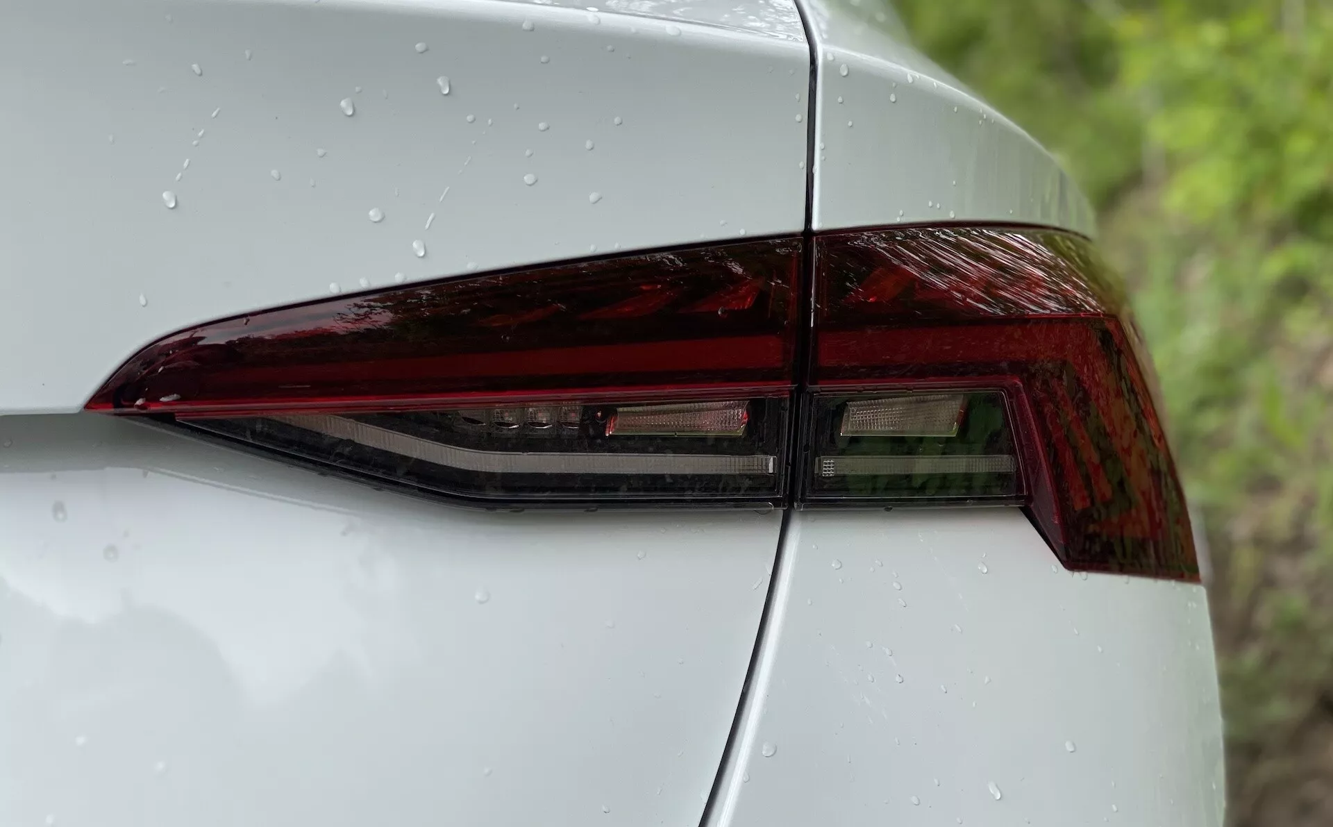 Närbild av vänster bakdel av en Škoda Octavia 2020 med fokus på bakljusenheten. Bilden är tagen från en diagonal vinkel som framhäver den moderna designen av LED-bakljuset med skarpa vinklar och en glansig finish. En del av bakre kvartspanelen och bagageluckan är synlig, vilket visar på en sömlös integration och aerodynamisk estetik. Den släta ytan har vattendroppar, vilket indikerar nylig nederbörd eller en färsk tvätt.