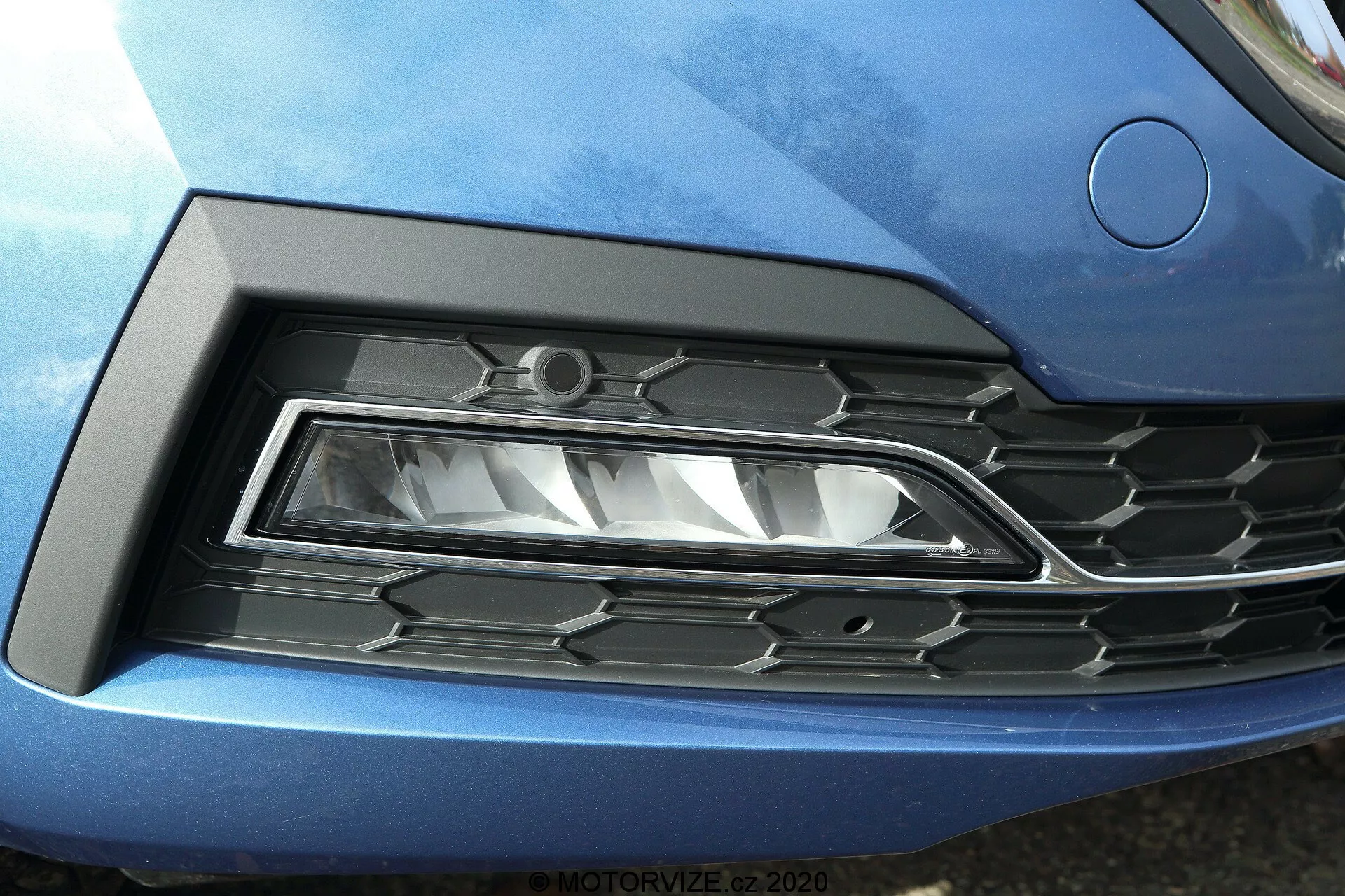Närbild av fronten på Skoda Octavia 2020 (Octavia Limousine), med fokus på vänster strålkastare och grill. Den moderna LED-strålkastaren med skarp, vinklad design är inplacerad i en skulpterad nisch som smälter sömlöst in i den metallicblå bilens kaross. Den intilliggande grillen har ett hexagonalt mönster av svart plast med krom-accenter, vilket återspeglar modern bilstyling.