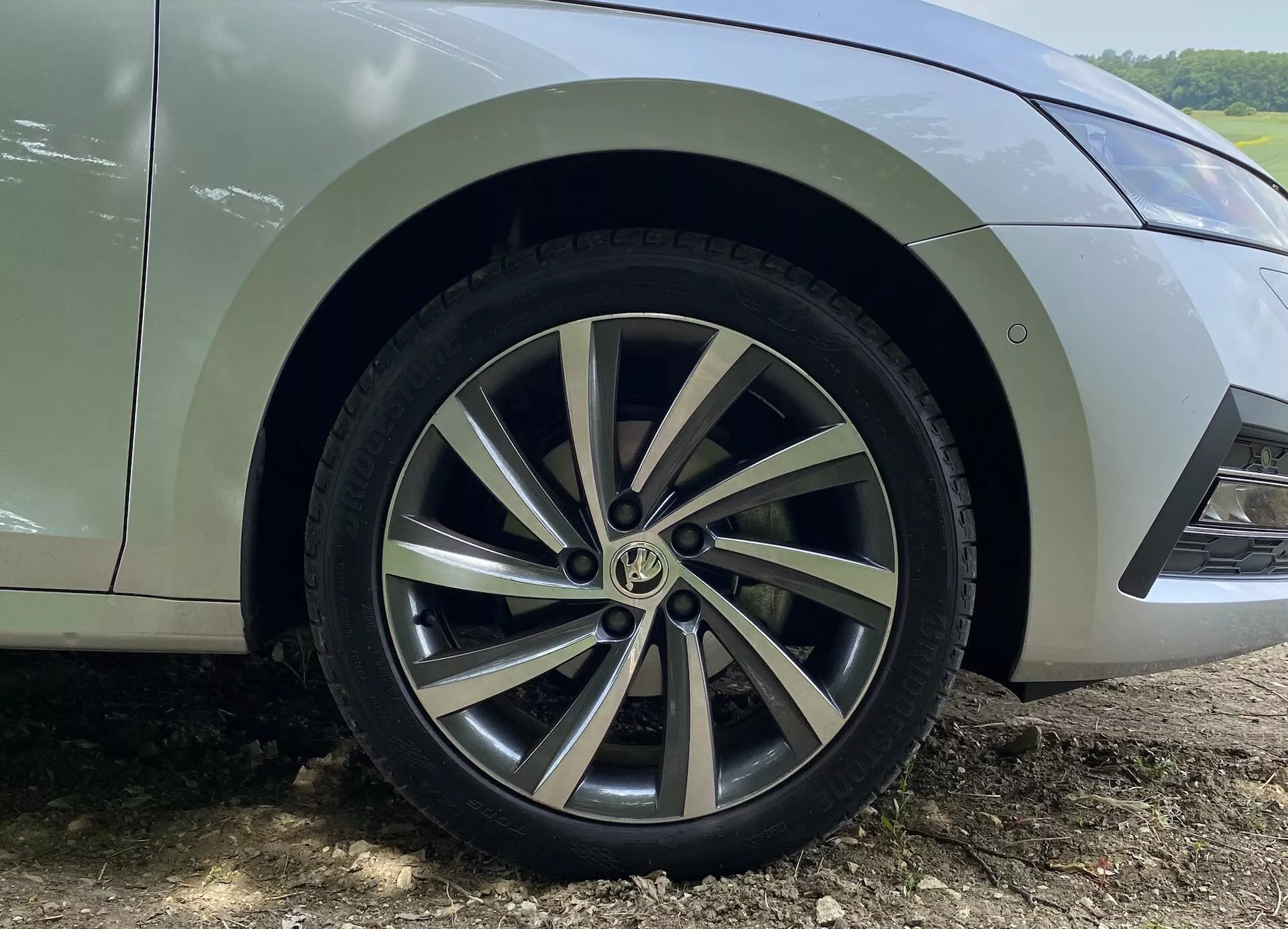 Närbild av framhjulet och den nedre stänkskärmen på en Skoda Octavia Limousine 2020, med fokus på den flerspåriga lättmetallfälgens design med en blandning av målad silverfärg och bearbetade ytor. Däckmönstret är synligt och visar lite slitage. Bilen är parkerad på en jordyta med synliga lerplask bakom hjulet, vilket tyder på senaste användning utanför vägen eller på en grusväg. Endast delen från den nedre främre dörren till frontbumpen är synlig från en låg vinkel, som fångar hjulets och stänkskärmens detaljer.
