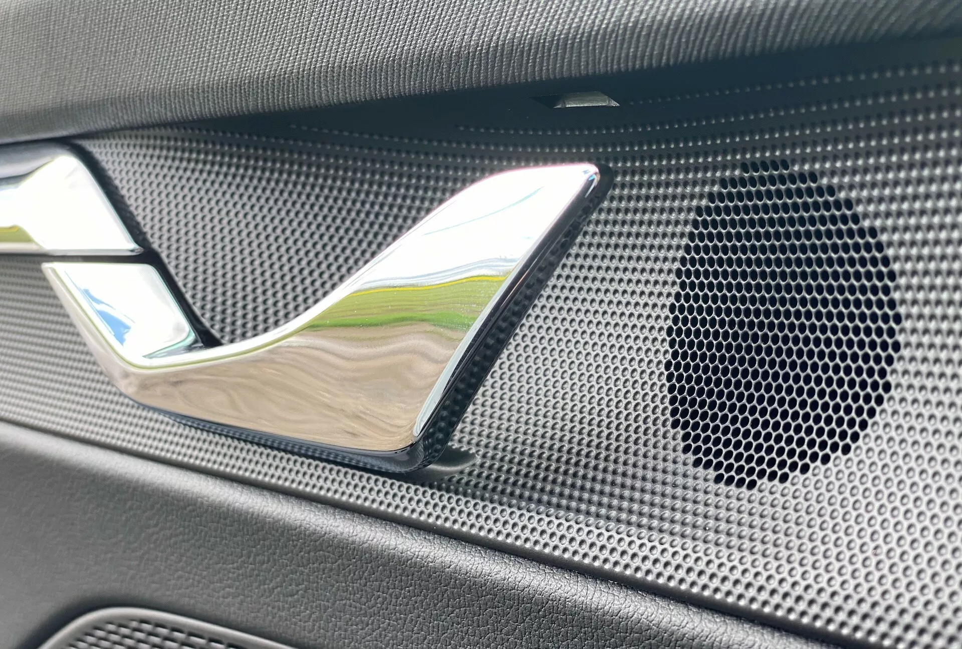 Interiördetaljbild av 2020 Skoda Octavia limousine dörrpanel, som framhäver dörrhandtaget i kromfinish och högtalargallret med ett bikakemönster i texturerat, eventuellt kolfibermaterial, vilket betonar lyx och högkvalitativt ljudsystem.