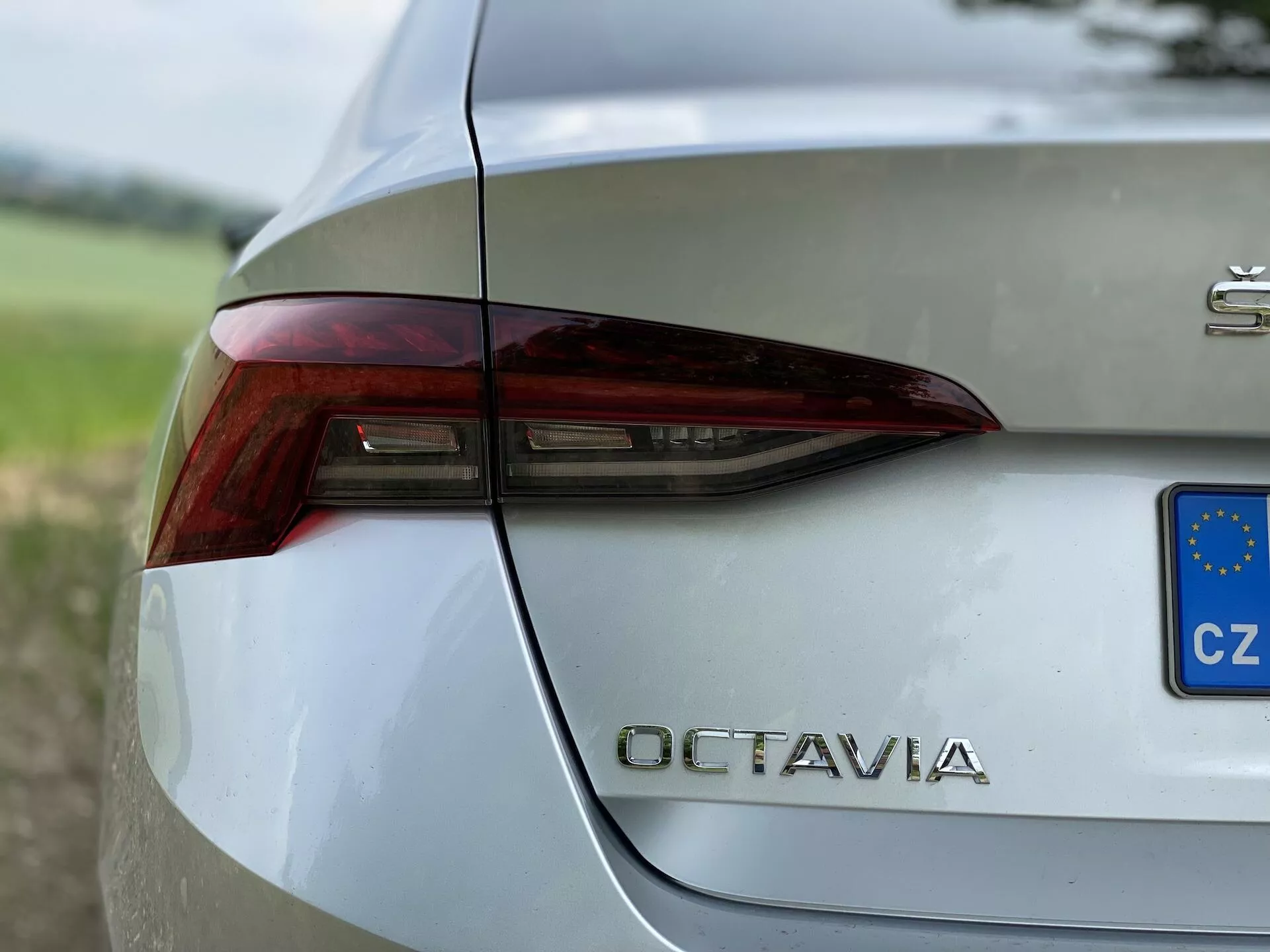 Närbild på bakre högra delen av en silverfärgad Škoda Octavia 2020. Bilden fångar den skarpa och kantiga designen av bakljuset med integrerade ljuselement för bromsljus, blinkers och eventuellt backljus. Bagageluckan visar tydligt modellbeteckningen 'OCTAVIA' i krombokstäver under bakljuset. En liten blå och gul rektangel med stjärnor indikerar registrering i Tjeckien. Fordonets polerade, metalliska yta reflekterar omgivningen och är ställd mot en suddig grön bakgrund, som framhäver dess sofistikerade design.