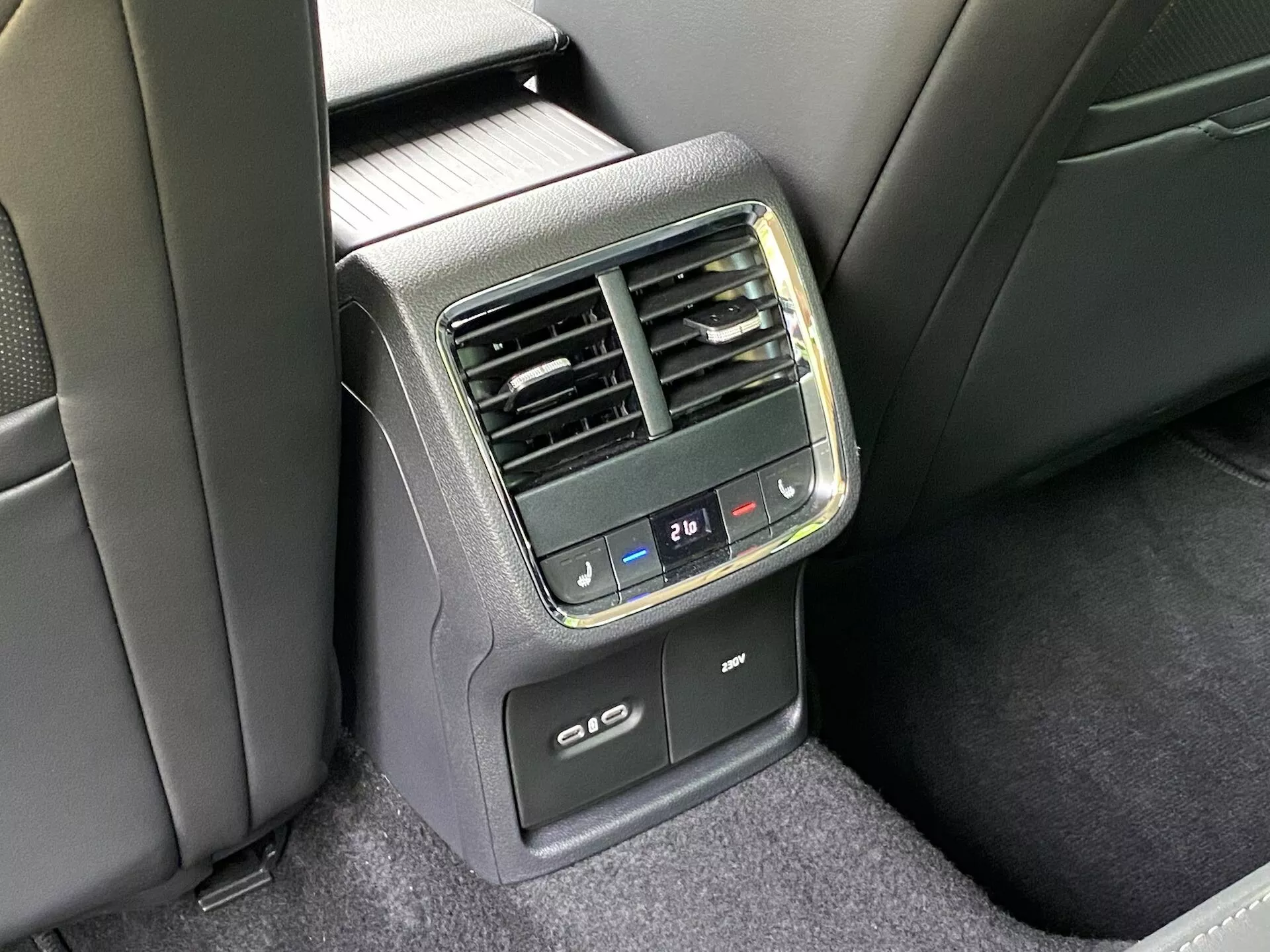 Detaljerad vy av den bakre klimatanläggningspanelen i baksätet på en Skoda Octavia limousine 2020. Tagen från en off-center vinkel, vilket ger en klar uppfattning om den centralt placerade panelen på baksidan av framsätets armstöd. Utrustad med digitala klimatanläggningsknappar för temperaturinställningar, vertikala luftventiler, en digital temperaturvisning inställd på 21℃, USB-laddningsportar och eventuell funktionalitet för uppvärmning eller kylning av baksätespassagerare. Materialen är en blandning av plast och tyg, i ett svart och mörkgrått färgschema under naturlig belysning.